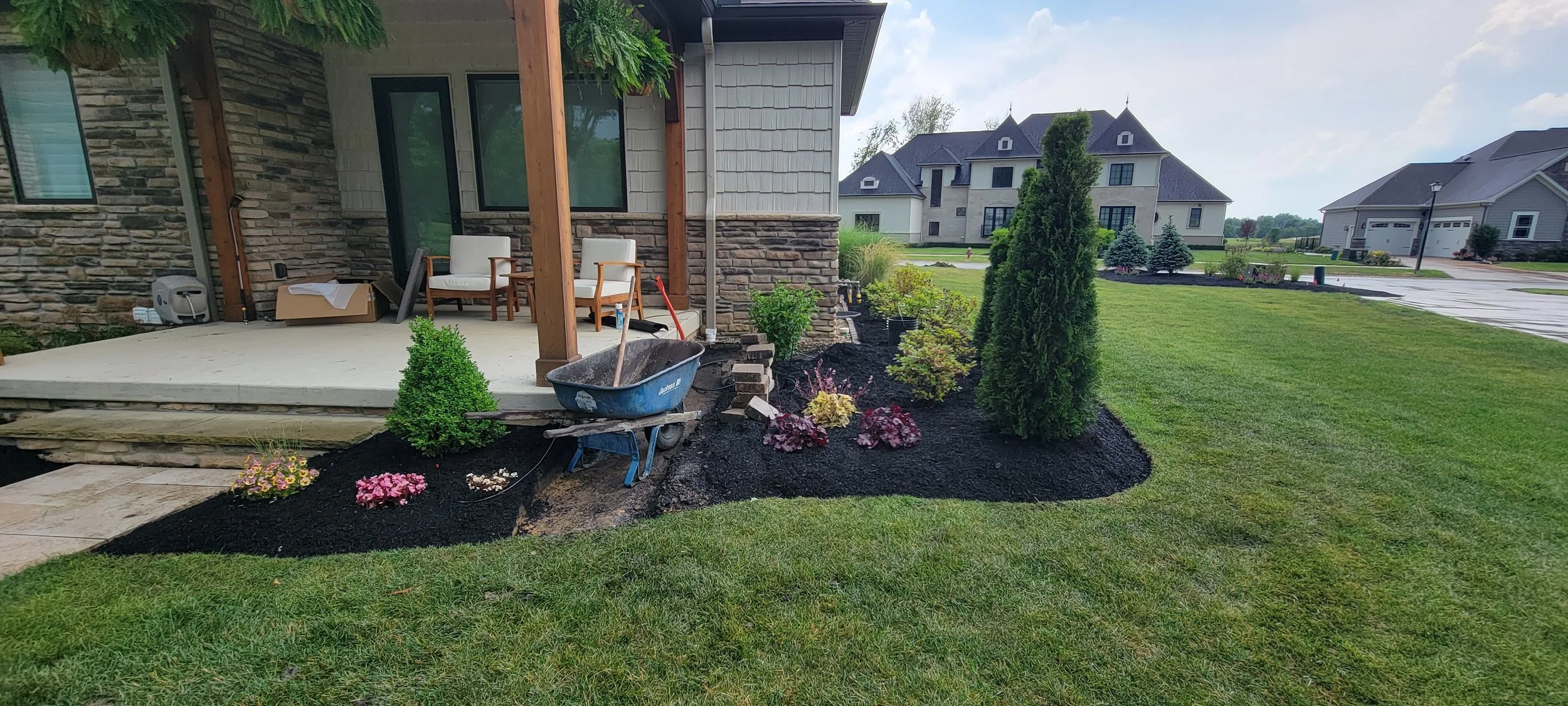 A backyard garden being landscaped with freshly planted flowers, shrubs, and small trees around a patio area. There are a blue wheelbarrow, a shovel, and some chairs on the patio, with nearby houses and a street in the background.