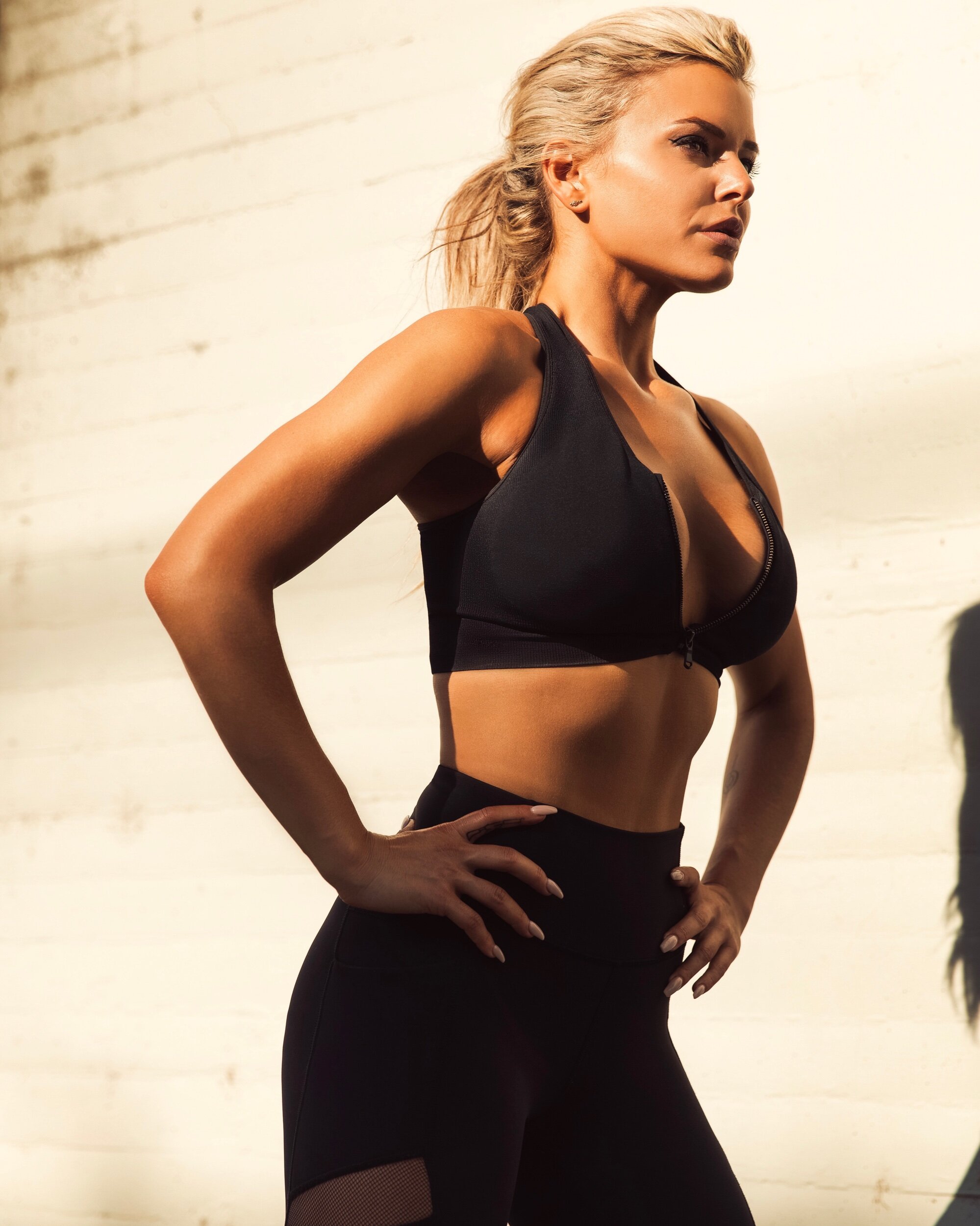 Female athlete wearing black workout gear, standing with her hands on her hips, in front of a light-colored wall.