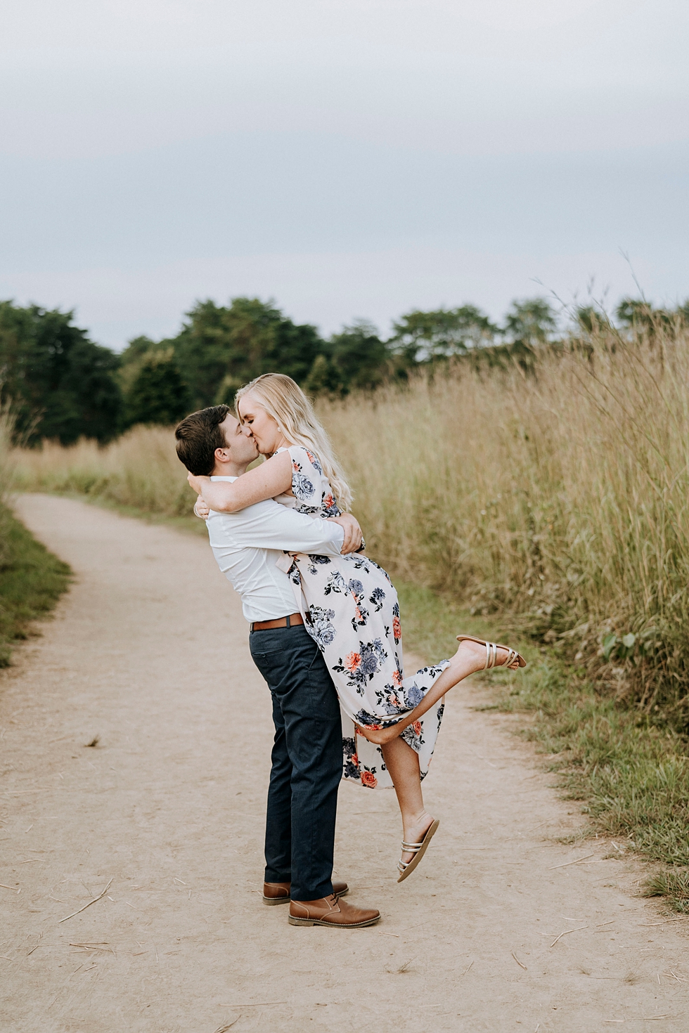  Field photo on forest pathway with couple kissing and bride to be wearing a floral dress with her leg popped #engagementphotographer #bernheimforest #engagementgoals #kentuckyengagementphotographer #kentuckyphotographer #engagementsession #photograp