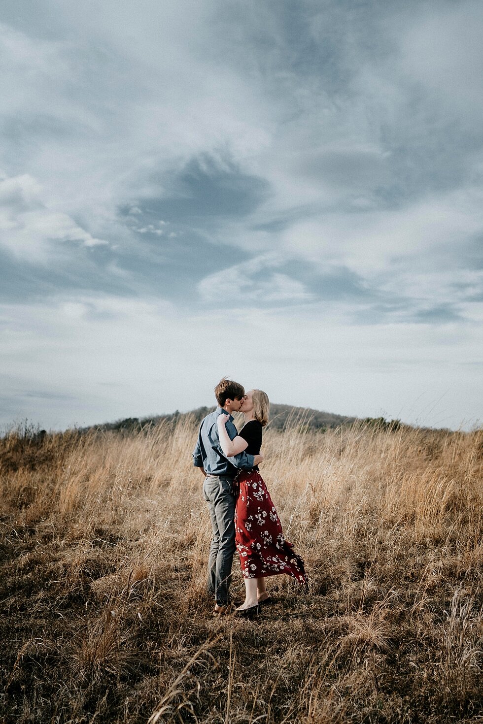  The beautiful contrast between the blue cloudy sky and the dry grass blowing in the wind was stunning for this Louisville, Kentucky engagement session. Kentucky mountains red floral skirt dark black shirt cloudy skies engagement session Photography 