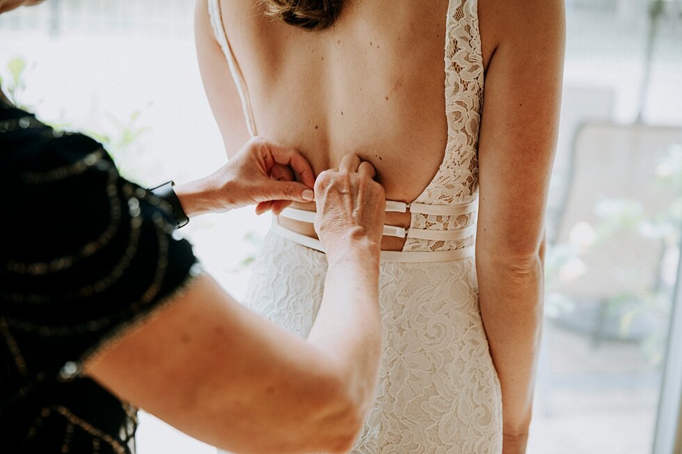  Bride’s mother helping her get ready for her wedding day as she clasps the back of her gorgeous wedding gown. the refinery jeffersonville indiana covid wedding outside wedding midwest louisville kentucky #weddingphoto #justmarried #midwestphotograph