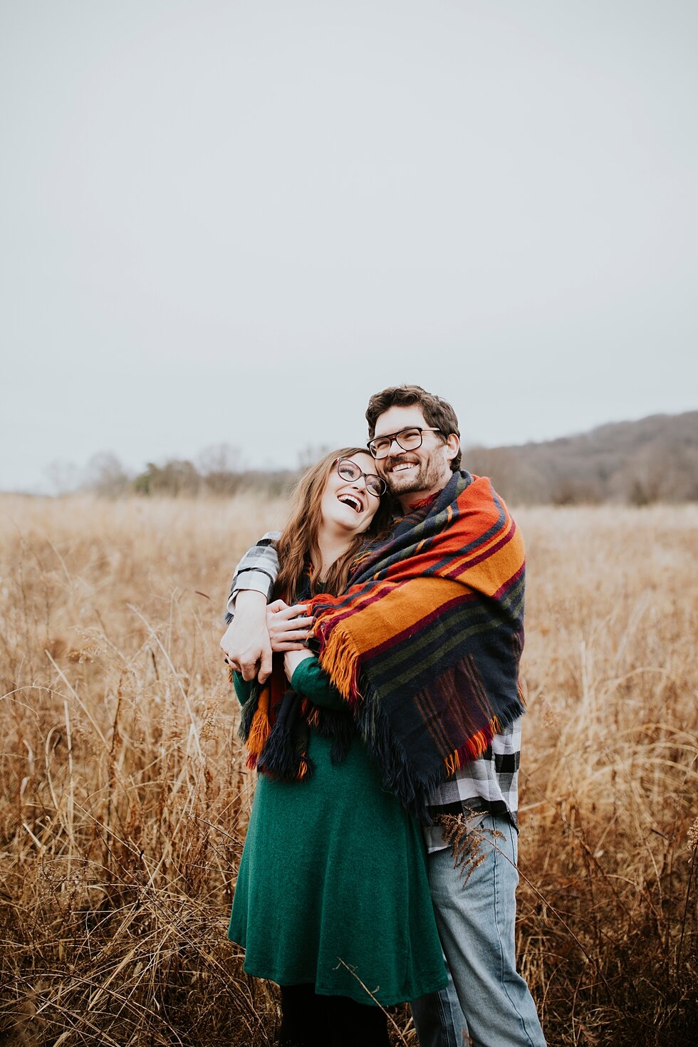  Engaged couple laughing under a warm orange and red blanket on a rainy winter day in Kentucky. Louisville photographer winter engagement Bernheim Forest emerald green dress plaid shirt rainy day Kentucky couple outdoor engagement  #savethedates #eng