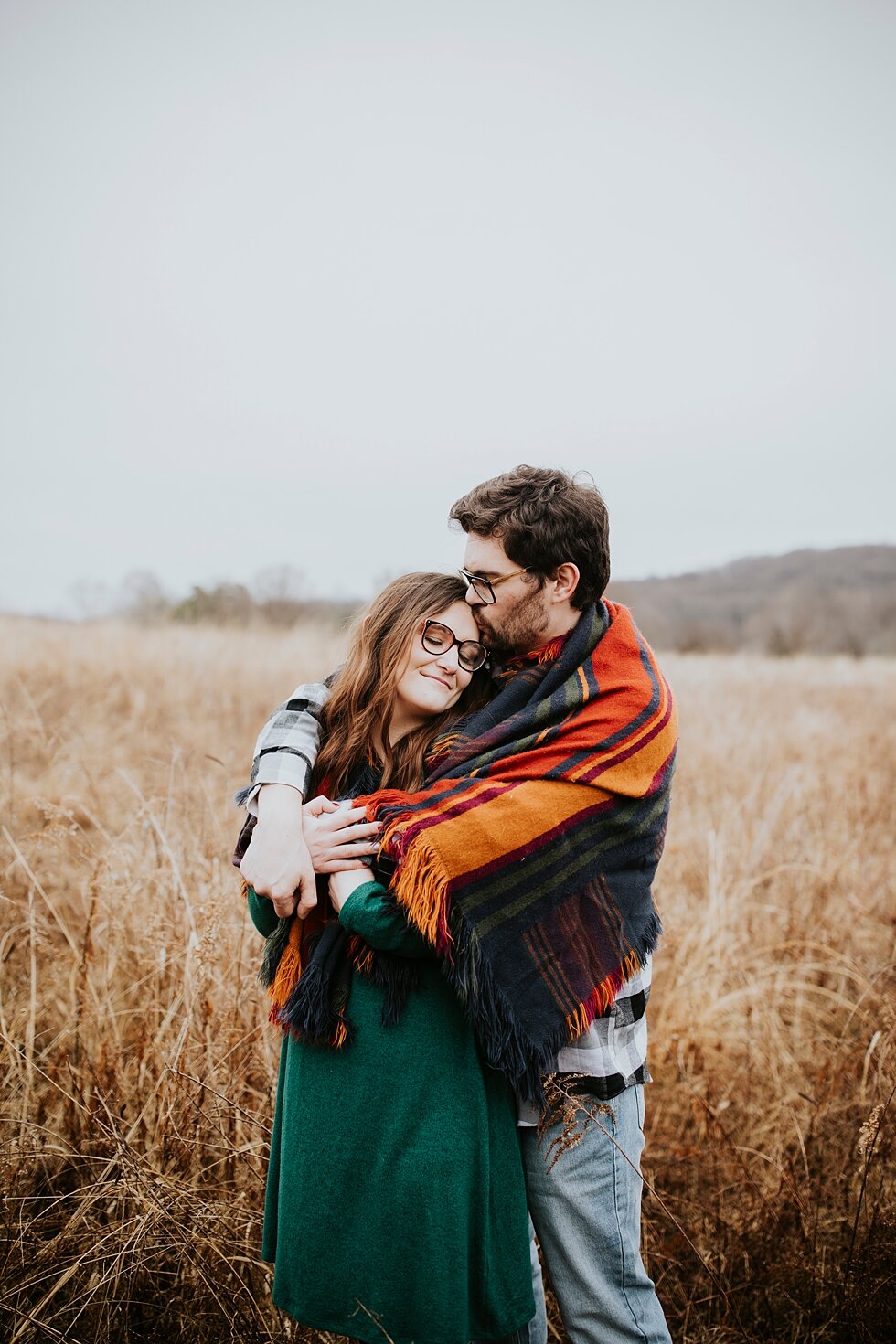  Sweet moments between man and woman as they huddle under a blanket on this rainy winter day in Louisville, Kentucky. Louisville photographer winter engagement Bernheim Forest emerald green dress plaid shirt rainy day Kentucky couple outdoor engageme