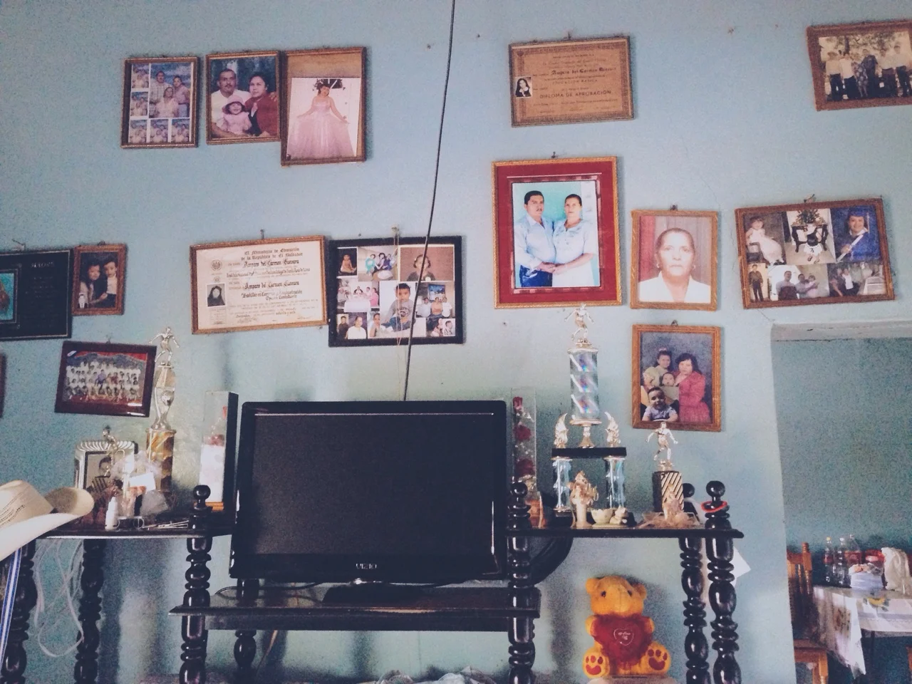 Cada hogar en este pulgarcito de este país tiene una pared cómo ésta. Fotos de los hijos grandes, nietos lejanos, padres fallecidos, títulos de bachillerato y retratos formales estremece la paredes como una muralla de familia. “Estas son la gente que hemos querido. Estos son nuestra familia,” declaran los cuadros juntos.
Every home in this pulgarcito of a country has a wall like this one. Pictures of grown children, distant grandchildren, deceased parents, high school degrees, and formal portraits scatter the plaster as a family mural. “These are the people we have loved. This is our family,” the frames proclaim in unison.