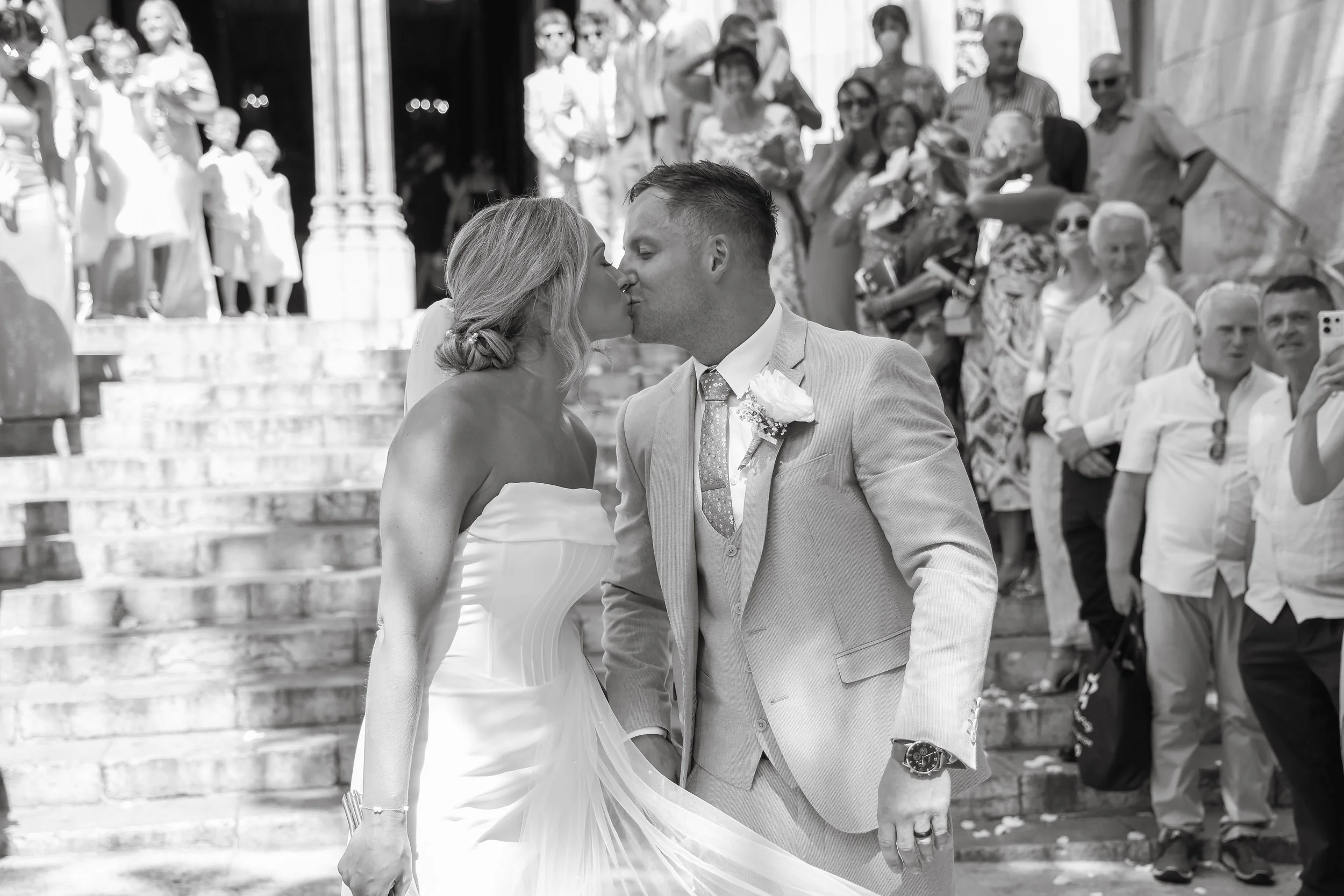 A bride and groom sharing a kiss on the steps outside a church during their wedding surrounded by friends and family