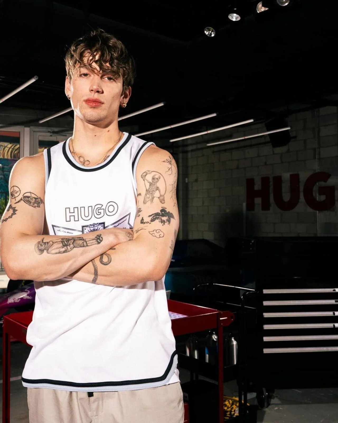 famous influencer Vic with tattoos, wearing a white basketball-style tank top with 'HUGO' logo, standing with arms crossed in a workshop or garage setting.