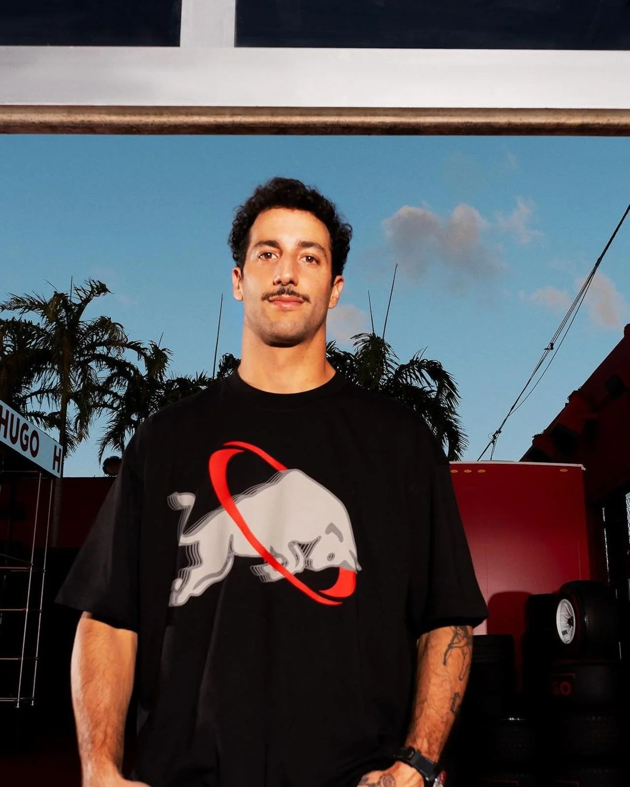 F1 Driver Daniel Ricciardo with dark curly hair, a mustache, and wearing a black T-shirt with a graphic of a dog and a red ring, standing outdoors against a backdrop of palm trees and some tires stacked nearby for HUGO BOSS MIAMI