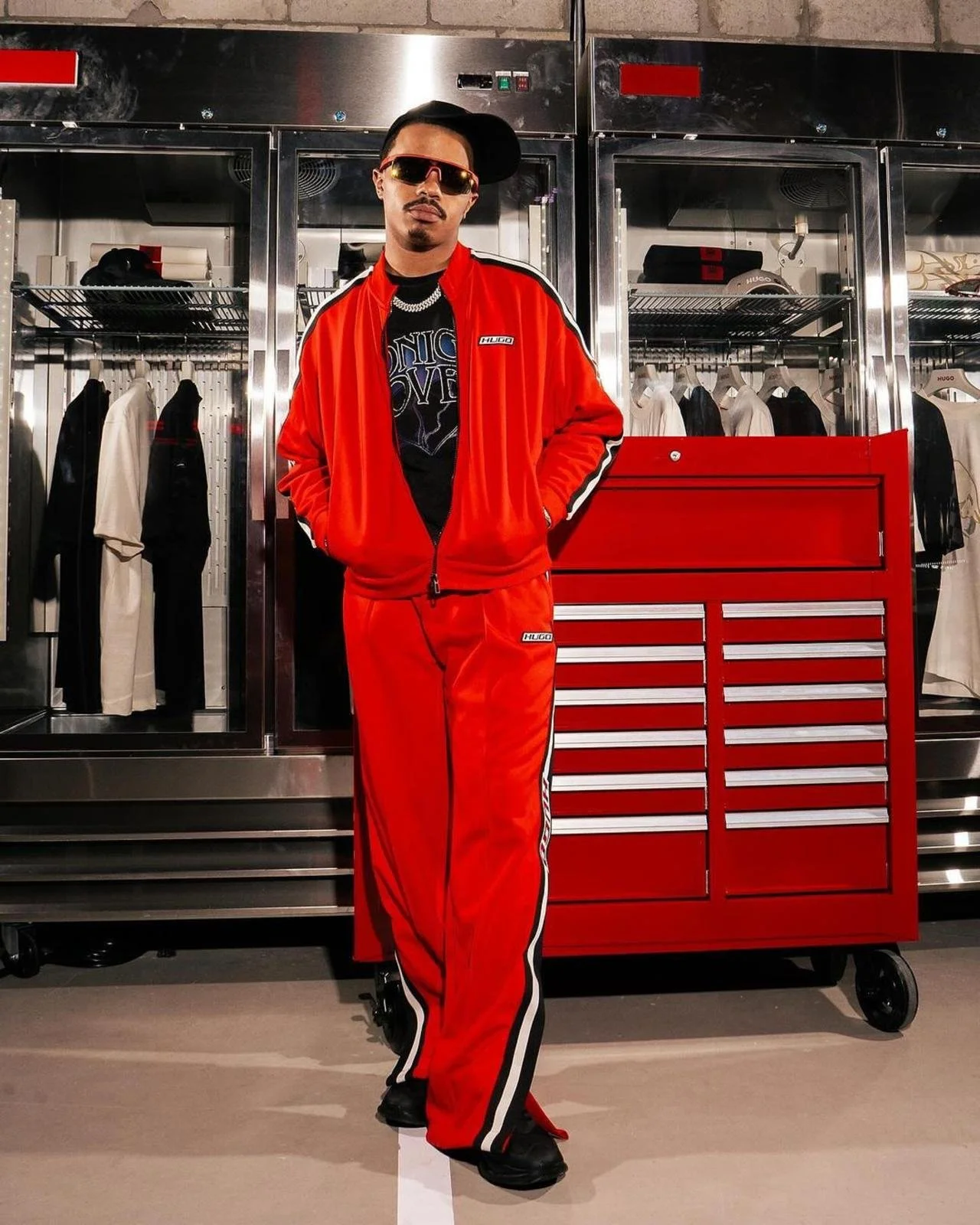 YG Marley in a red track suit with black and white stripes, black shoes, sunglasses, and a black cap tilted sideways, standing with hands in pockets in front of a red tool chest and chrome closet with clothing inside for Hugo Boss and f1 Miami popup