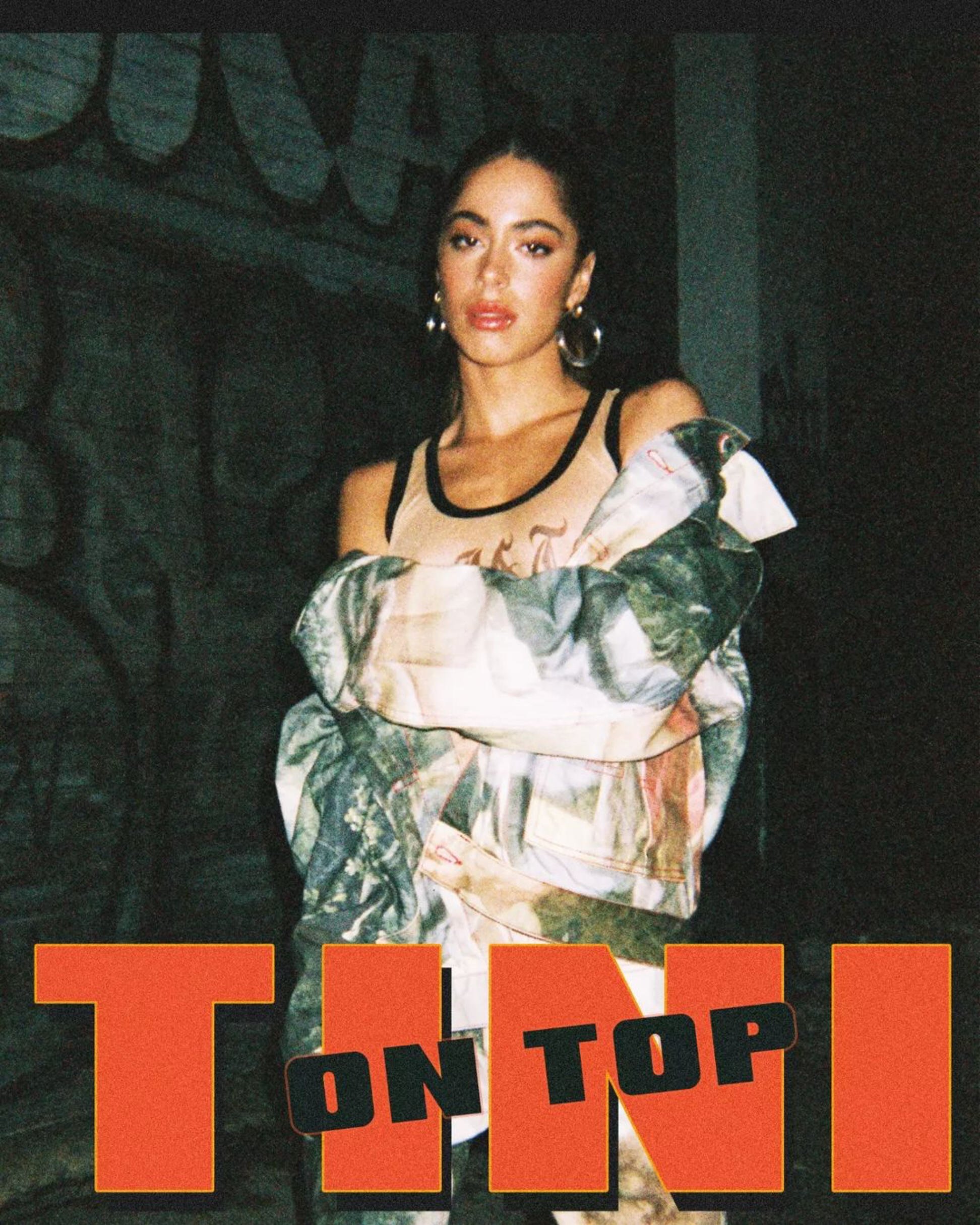 popstar TINI standing with arms crossed, wearing a tank top and a colorful jacket draped over her shoulders, with the text "TINI" and "ON TOP" in bold letters at the bottom of the image.