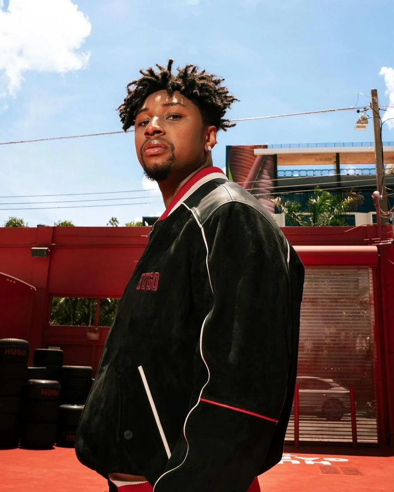 A young black man with curly hair and earrings standing outdoors on a sunny day, wearing a black jacket with red and white accents, in front of a red structure with tires and a building in the background.