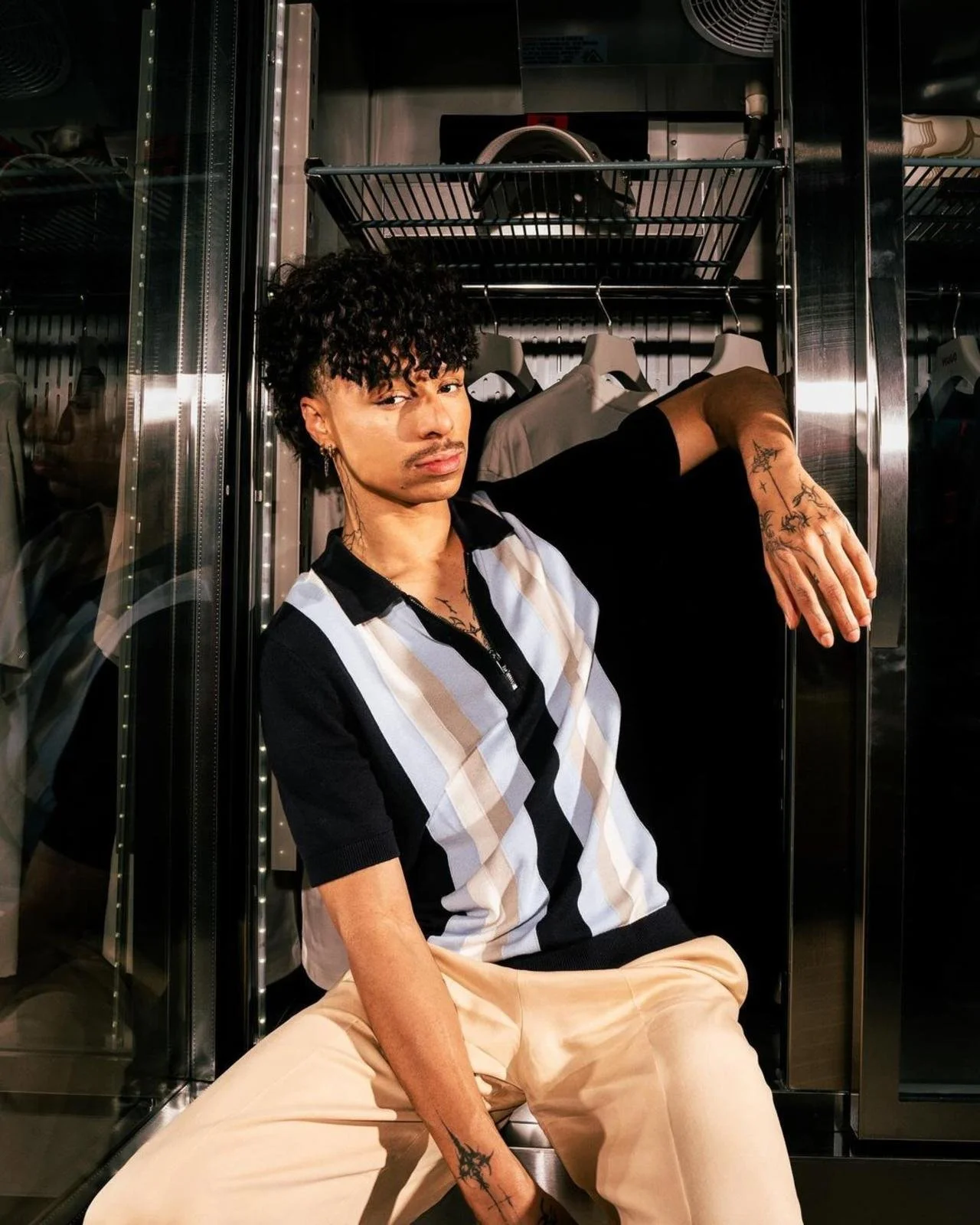 A person with curly hair, tattoos on their hand and neck, wearing a black, white, and beige striped shirt and beige pants, sitting in front of a metal storage cabinet with hangers and clothing in the background.