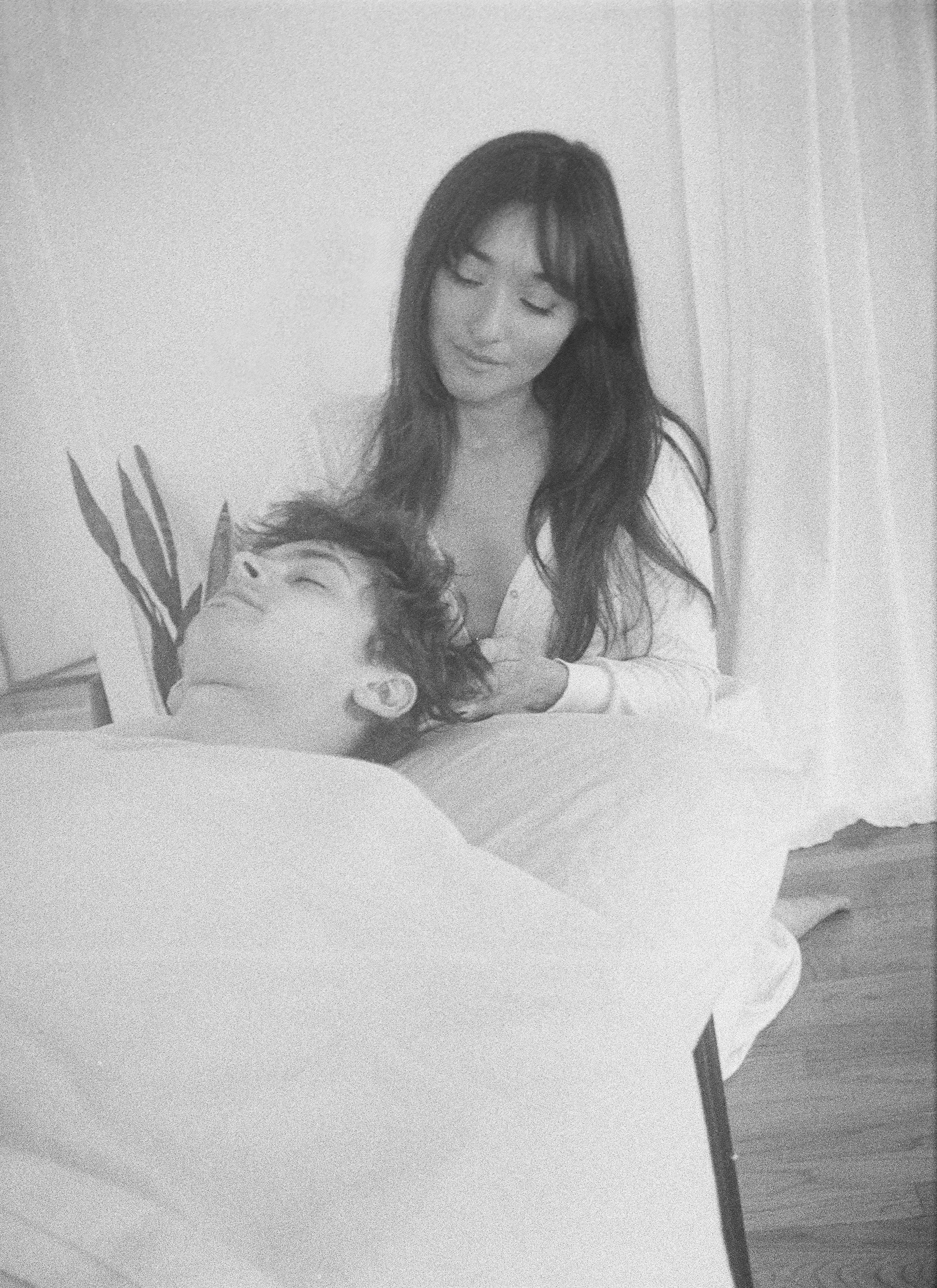 A woman looks down at a man lying in bed with a gentle smile. The man is resting with his eyes closed and appears content.