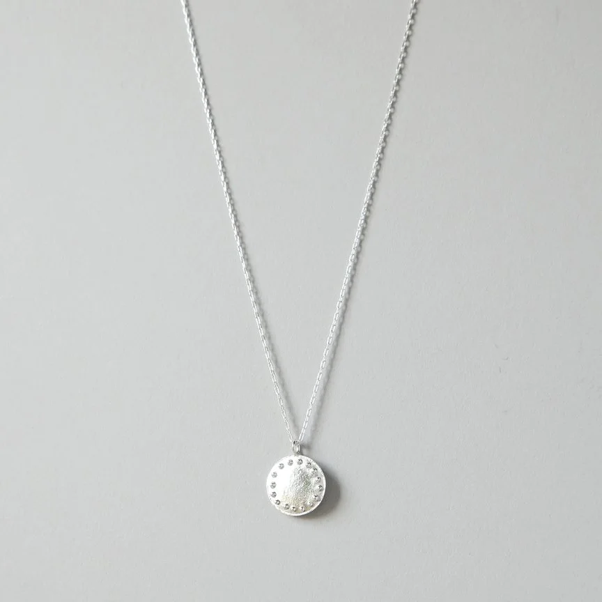 Image of a circle sterling silver elipse moon sun pendant necklace with granules on delicate chain front view on a gray blue background, minimal, simple and versatile