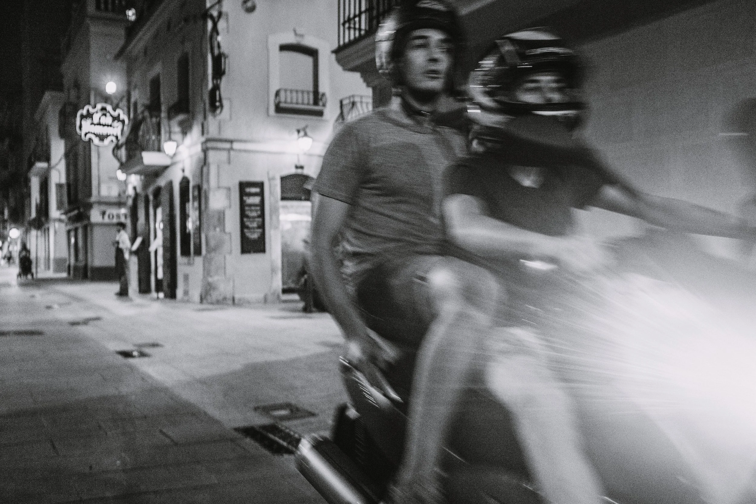 A young man riding a scooter at night on a city street, with buildings and streetlights in the background, in black and white.