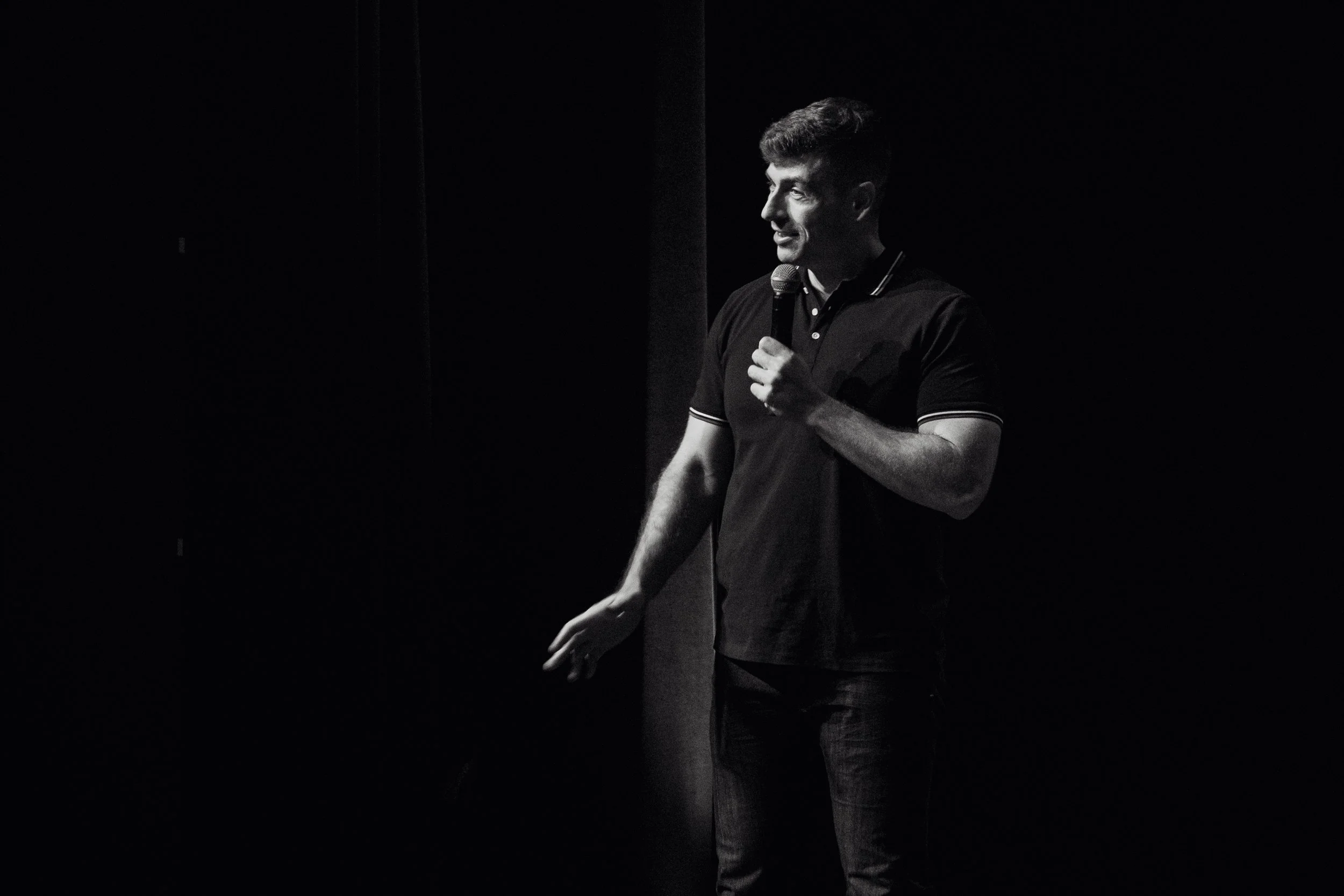 A man speaking into a microphone on stage, illuminated against a dark background.