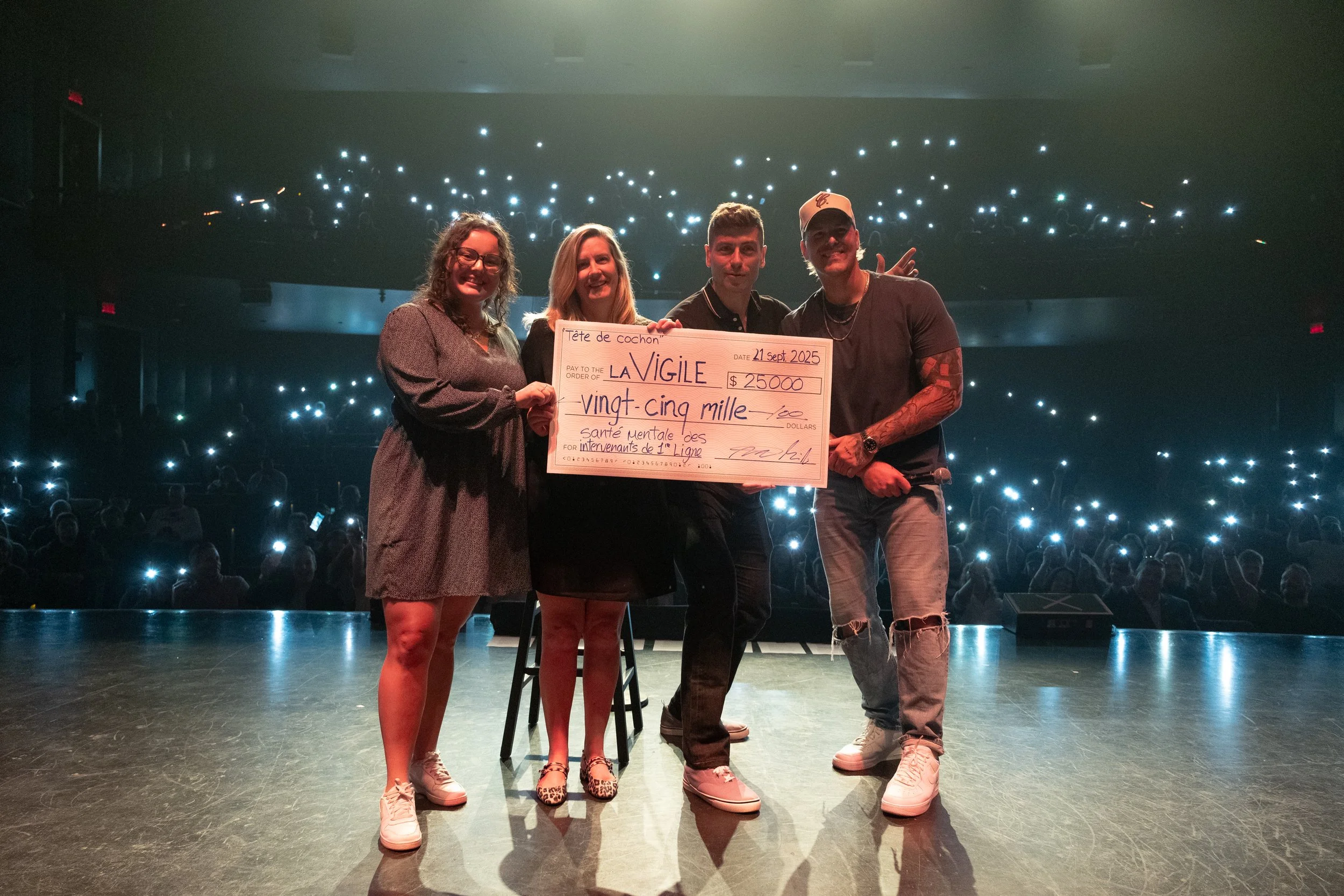Four people on a stage holding a large ceremonial check with an audience in the background. Stage lighting creates a bright and celebratory atmosphere.