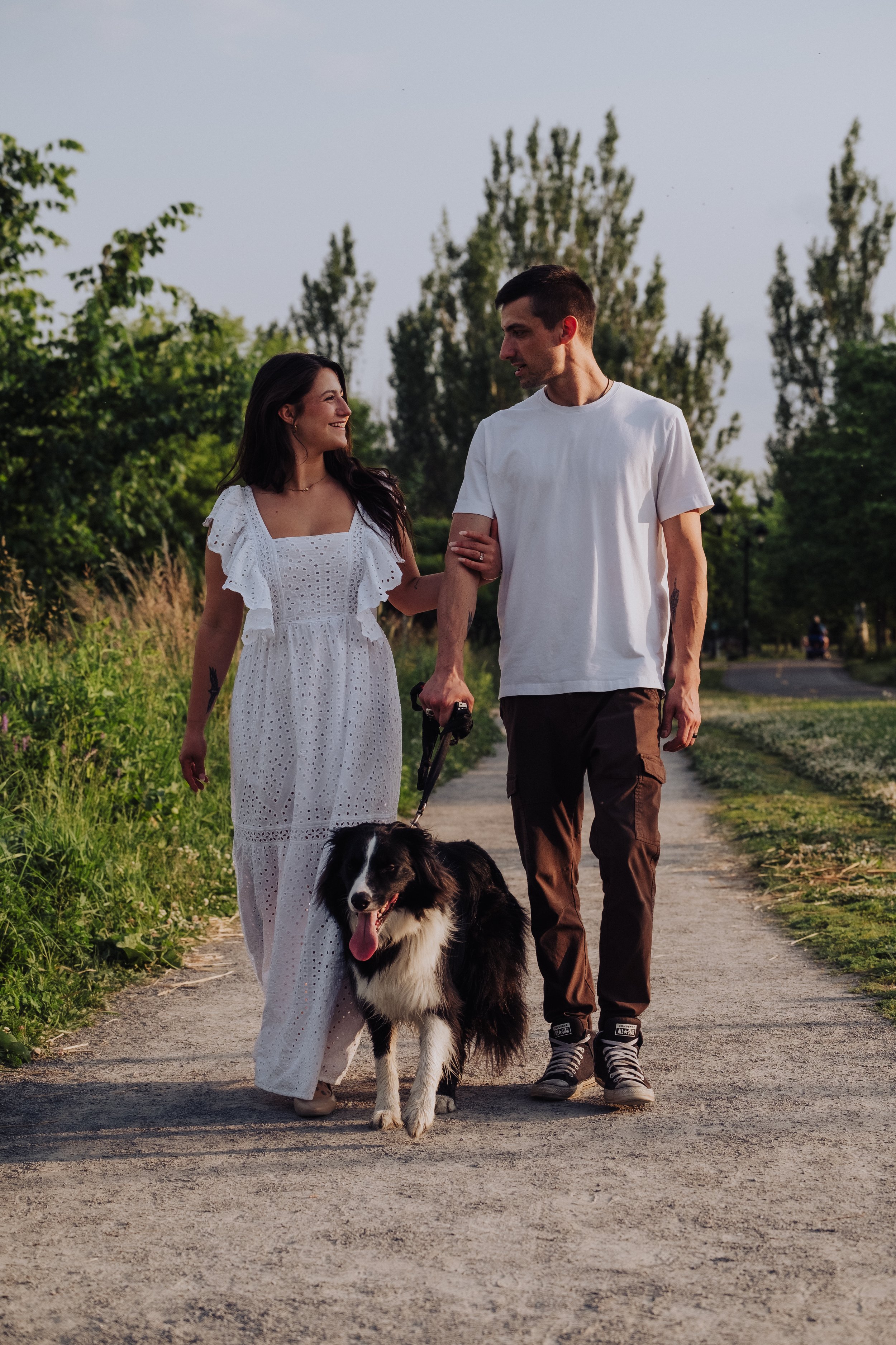 A young couple walking on a dirt path outdoors with a dog. The woman is wearing a white dress, and the man is in a white t-shirt and brown pants. They are holding hands and smiling at each other while the dog walks between them.
