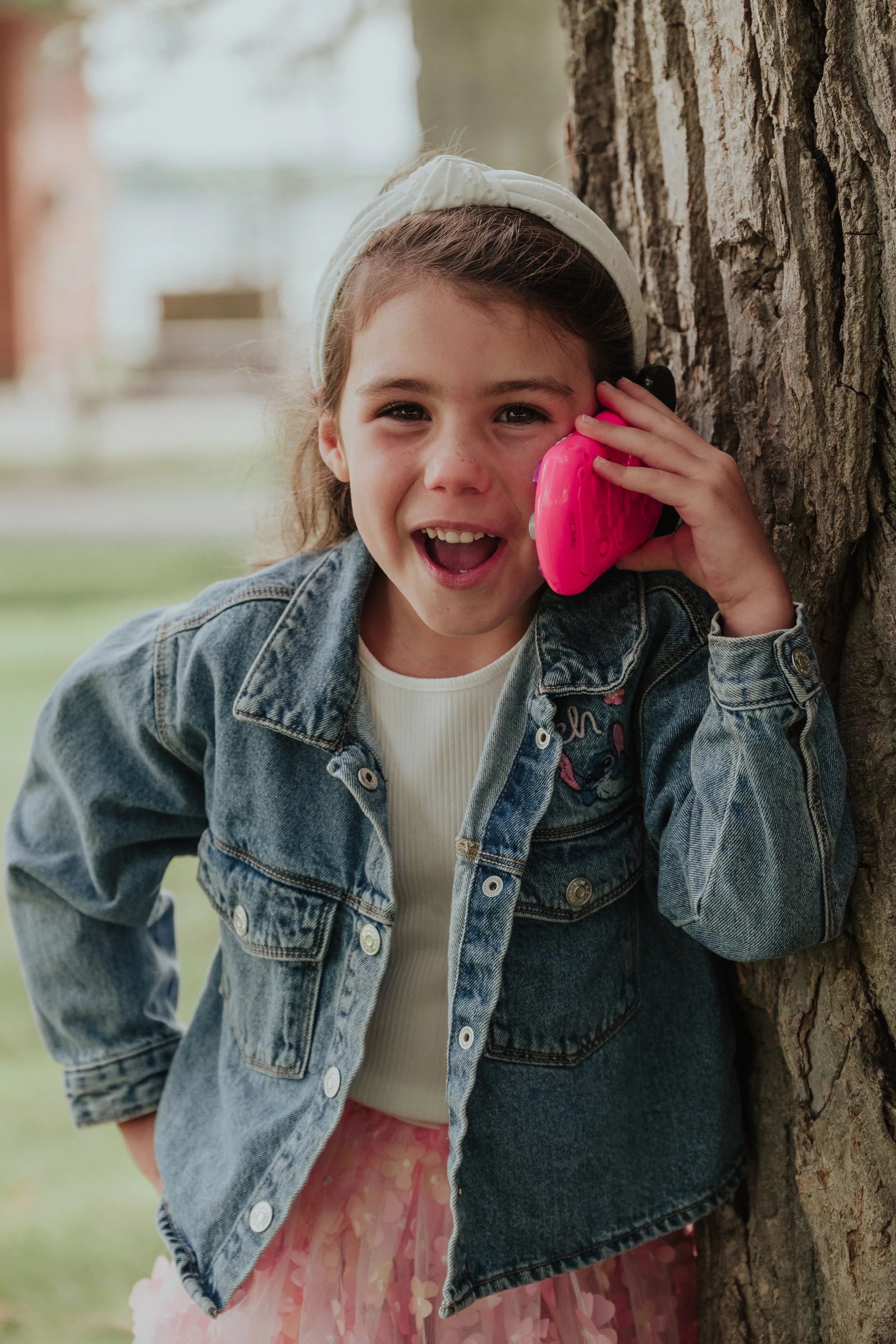 A young girl with a denim jacket and pink skirt talking on a pink toy phone outside by a tree.