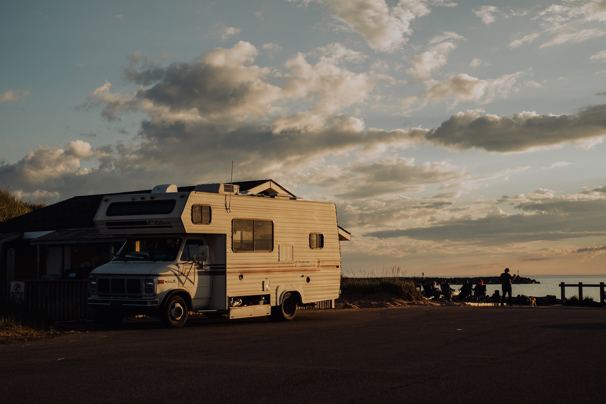 A recreational vehicle (RV) parked near a house during sunset, with people and dogs near the ocean, clouds overhead, and a fence along the shoreline.