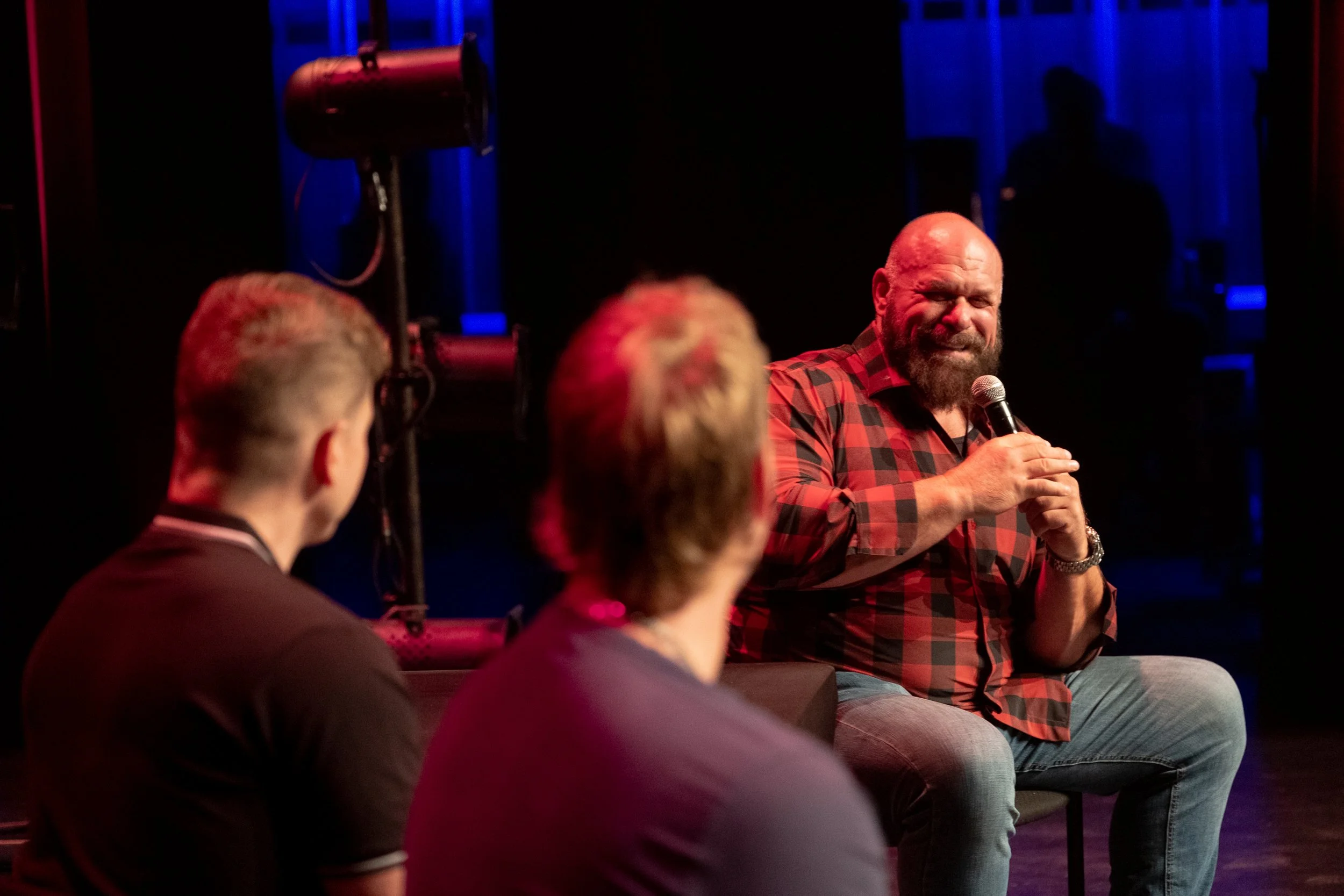 A man with a beard and bald head wearing a red and black plaid shirt sitting on a stage with a microphone, speaking to an audience. Two other people are seated in the foreground, listening.
