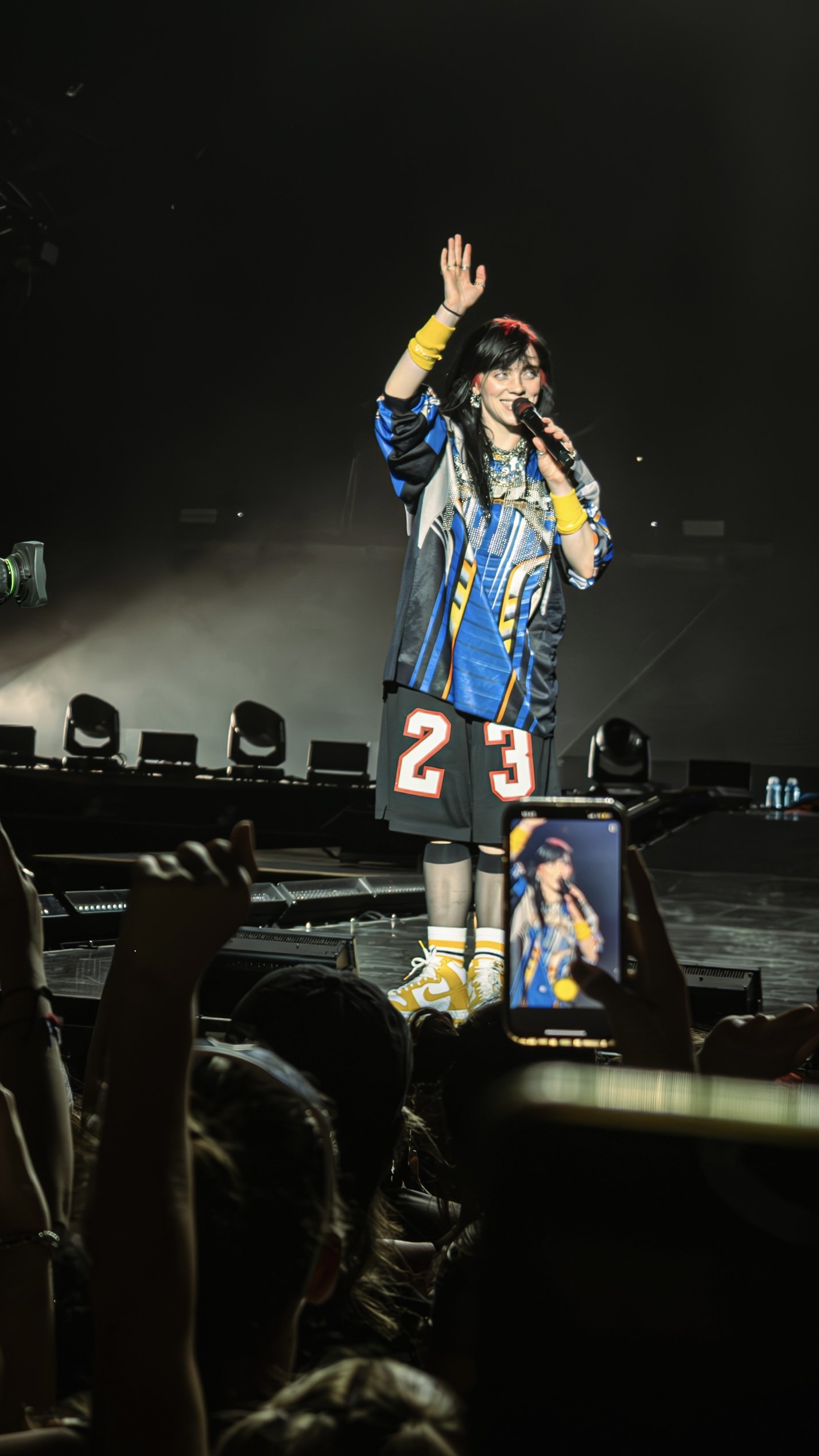 A woman on stage performing at a concert, holding a microphone and waving at the audience, with fans taking photos and videos.