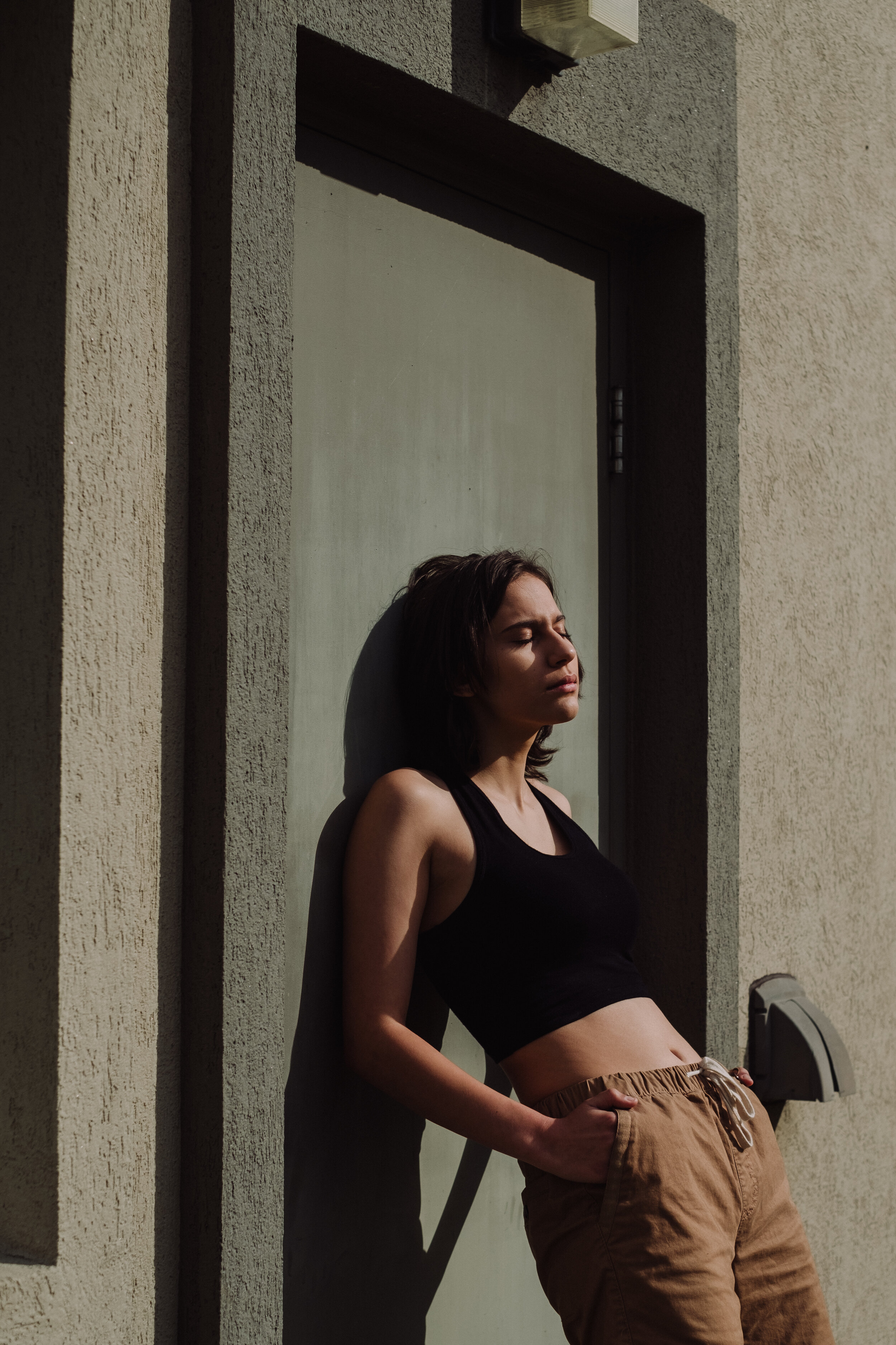 A woman in a black crop top and brown sweatpants leaning against a wall with her eyes closed on a sunny day.
