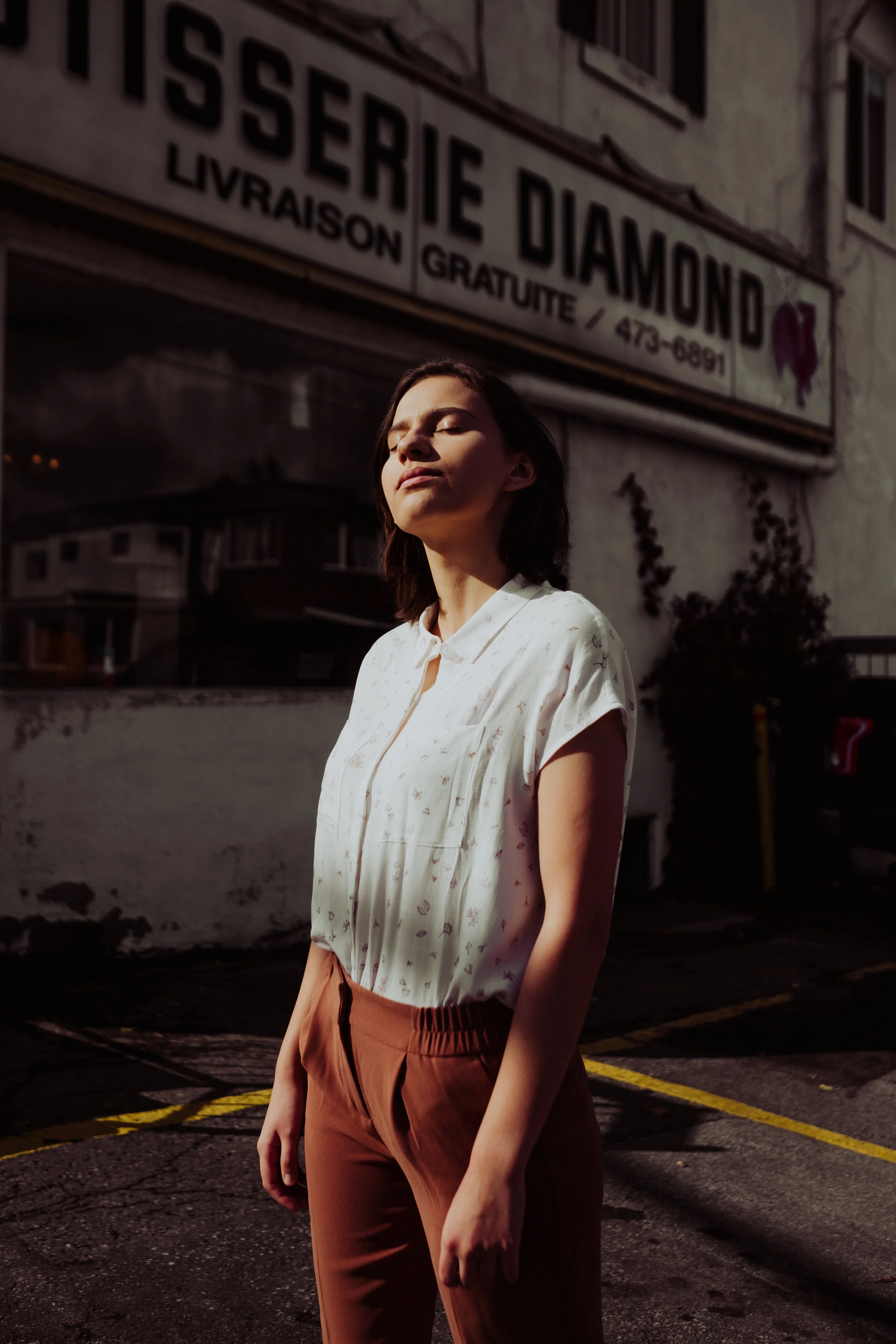 A woman standing outdoors with eyes closed and face tilted up, sunlit, with an 'Issérie Diamond' sign on a building in the background.