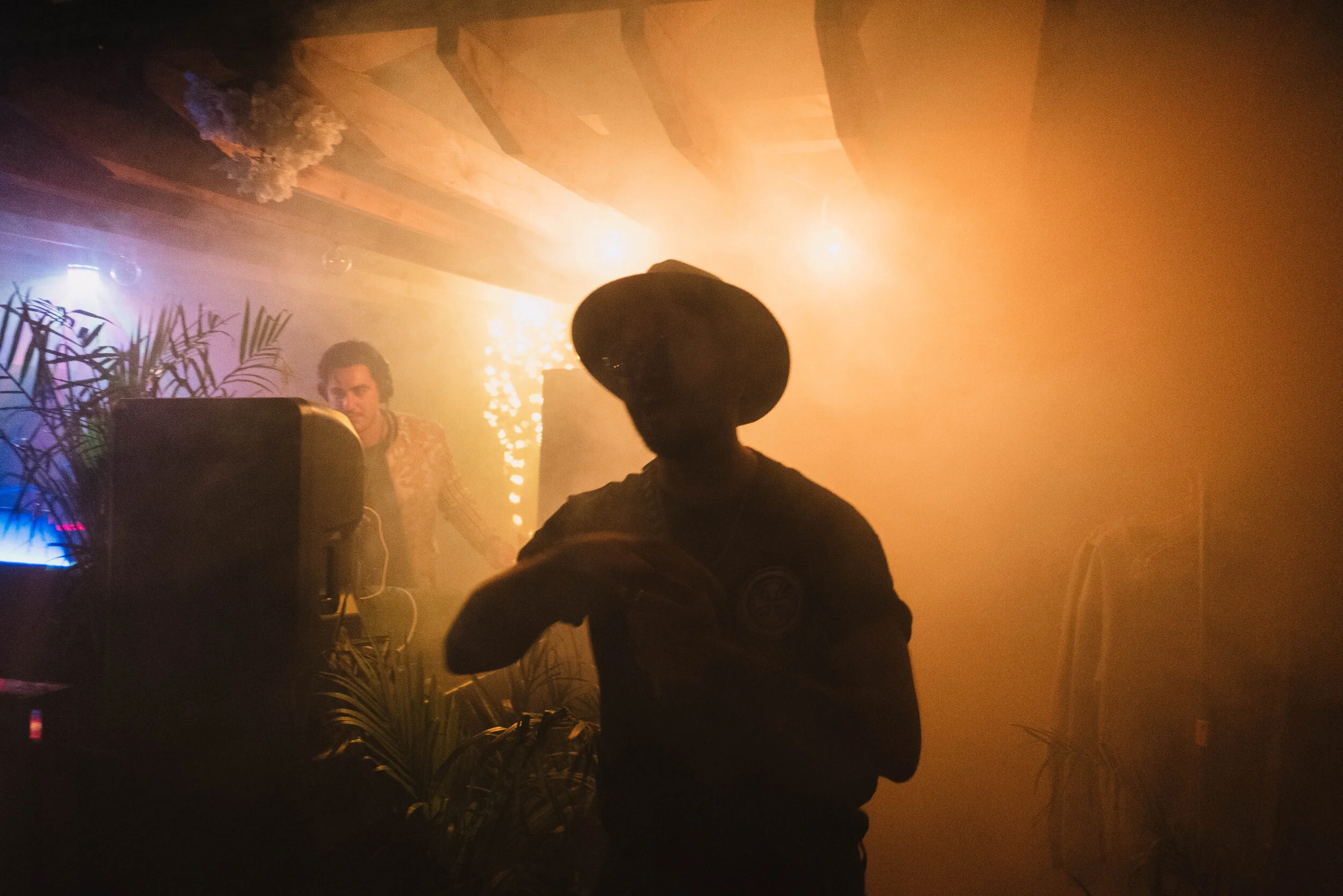 Silhouette of a person wearing a hat and a badge, with a DJ in the background playing music in a dimly lit, smoky nightclub with colorful lights.