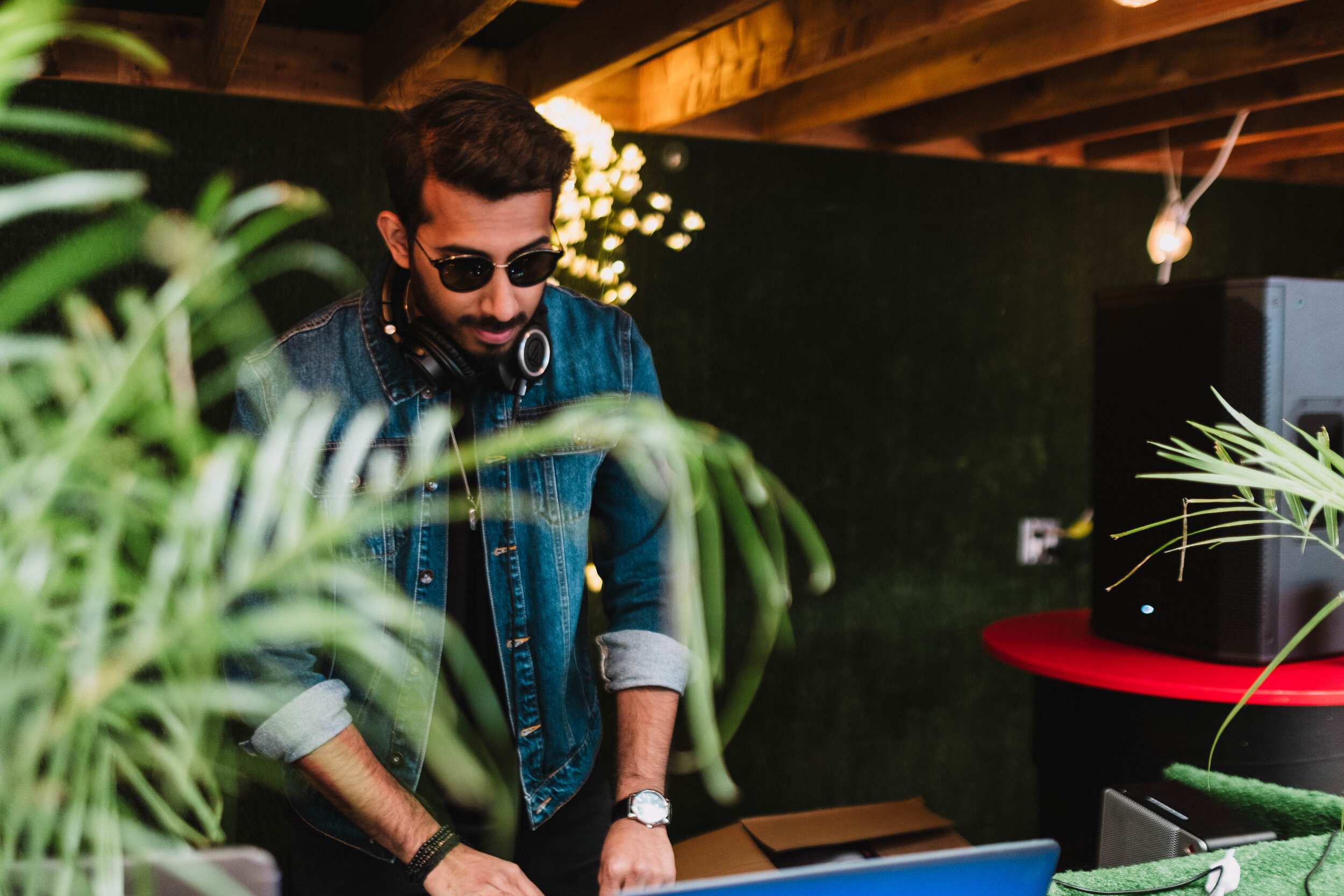 A man with sunglasses and headphones around his neck, wearing a denim jacket, is standing behind a laptop in a room with dark walls, plants, and warm lighting.