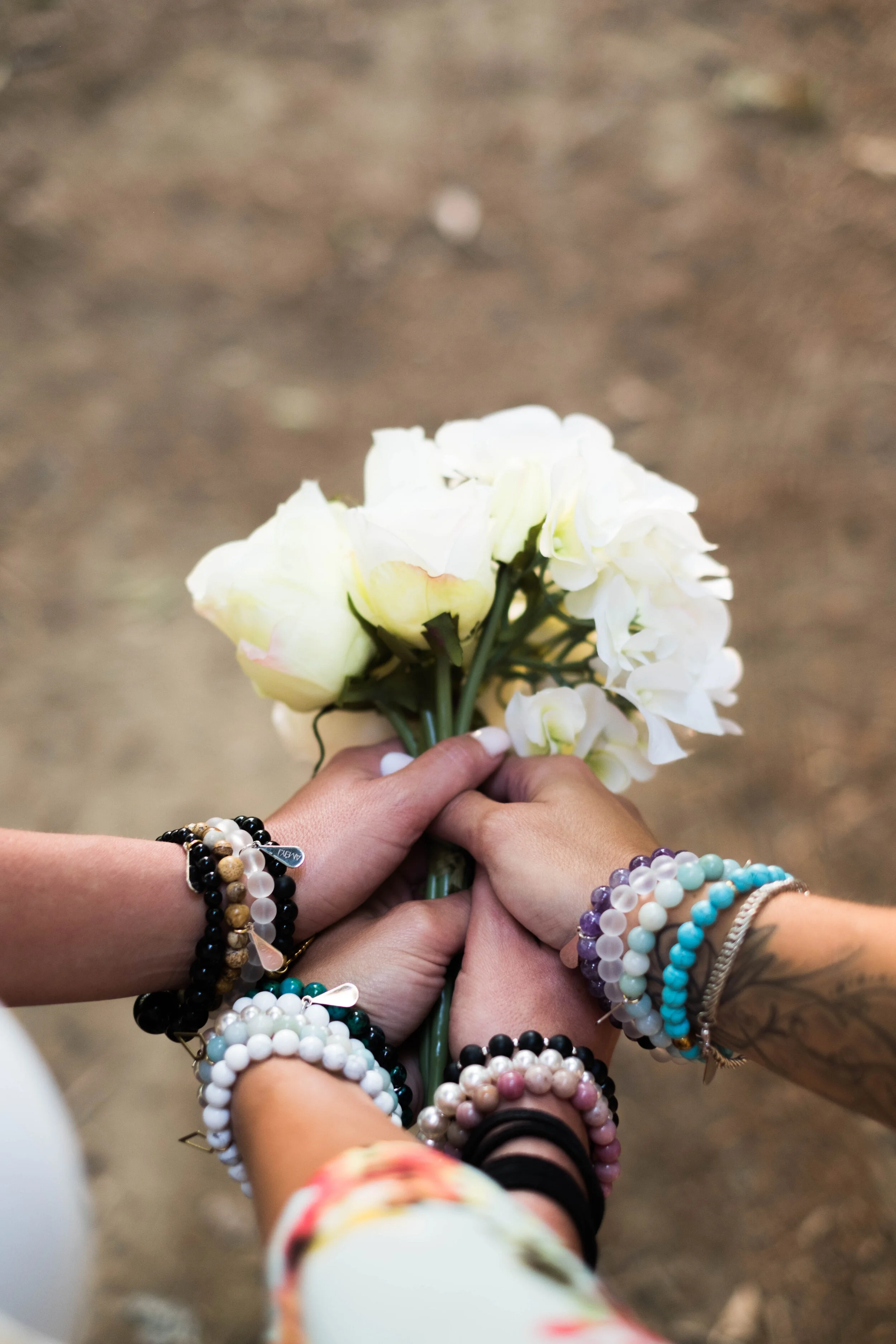 Multiple hands holding a bouquet of white flowers, adorned with various colorful bracelets.
