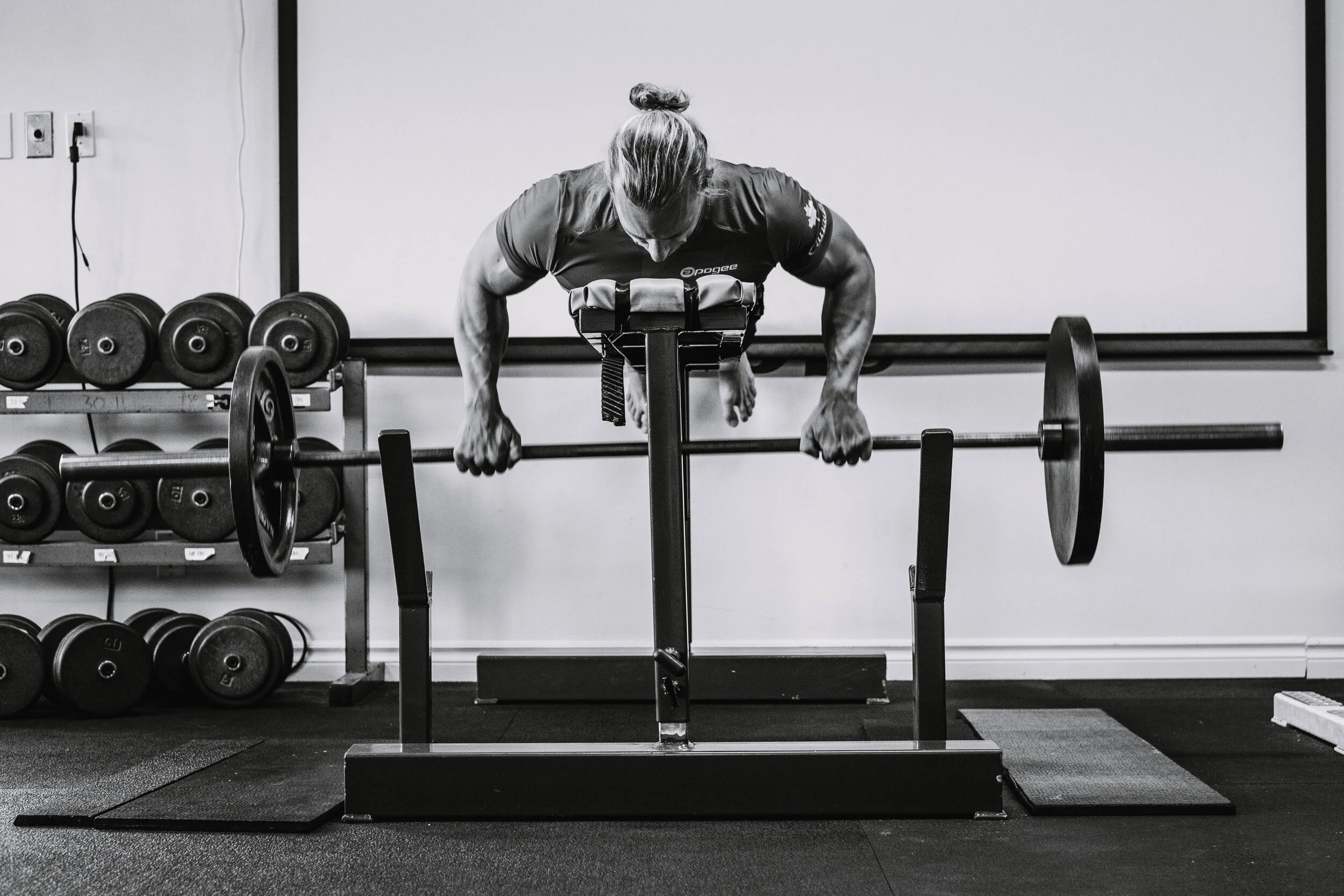 A woman in athletic clothing performing a push-up with a barbell on a squat rack in a gym.