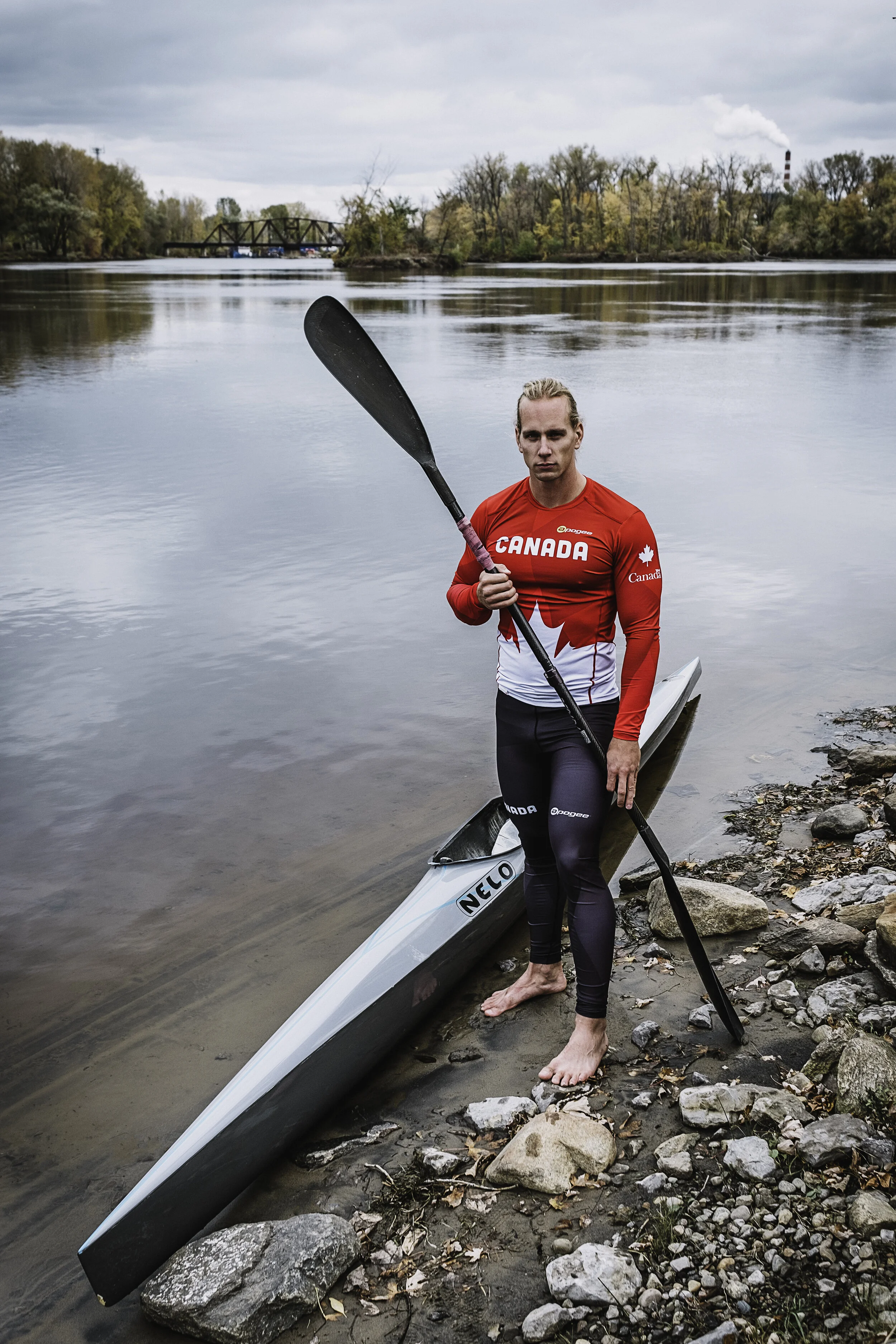 A person standing on a rocky riverbank holding a paddle next to a kayak, wearing a red Canada sports jersey and black leggings, with a calm river and trees in the background.