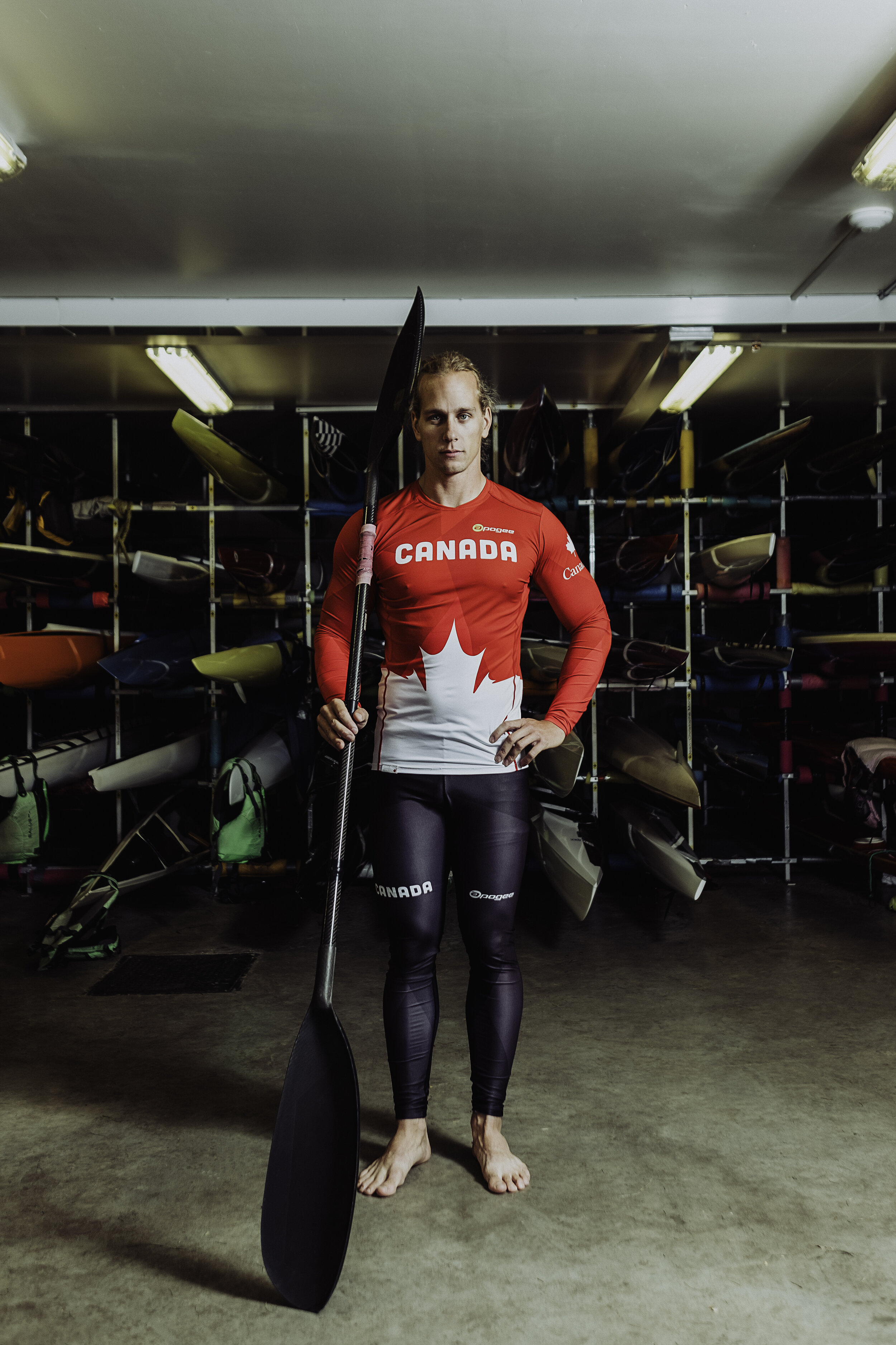 A female athlete with long hair tied back, standing barefoot indoors in front of shelves filled with kayaks and paddles. She is wearing a red and white Canada sports uniform and holding a paddle, looking directly at the camera.