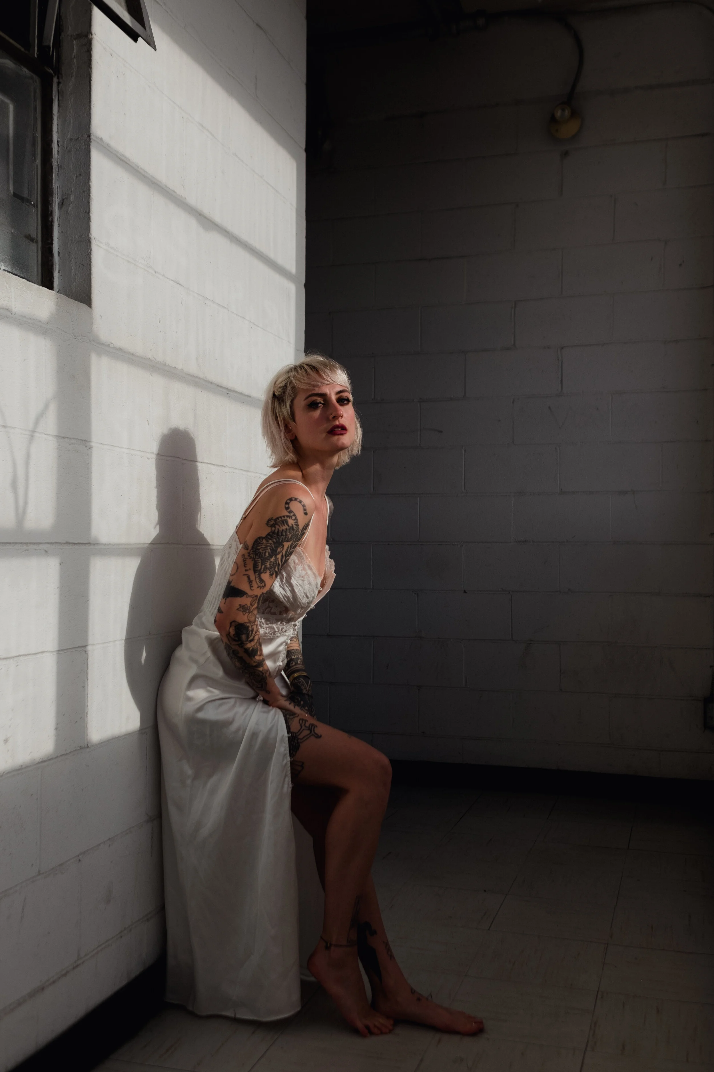A woman with blonde hair and tattoos sitting on the floor in a dimly lit room, wearing a white dress.