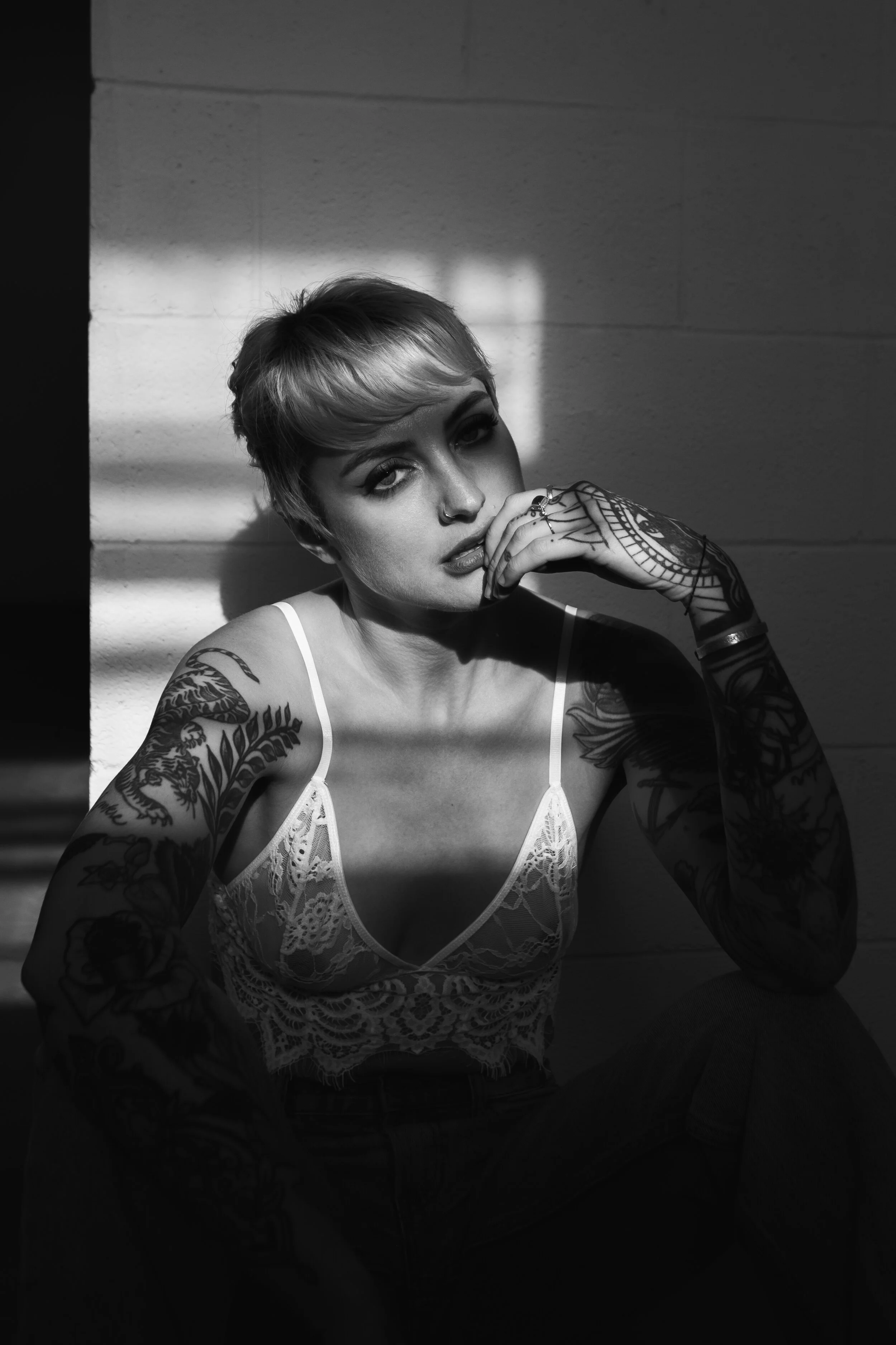 A black and white photo of a tattooed woman with short hair, wearing a lace top, sitting against a wall with shadows cast across her face, looking at the camera.