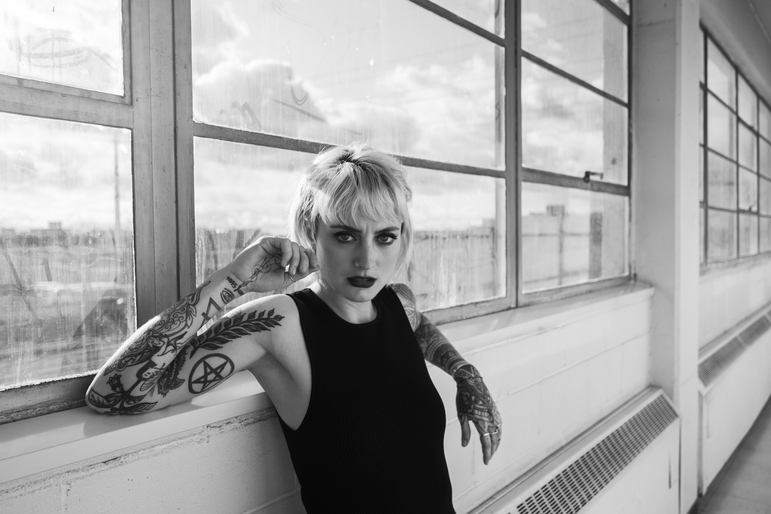 Black and white photo of a woman with short blonde hair and tattoos on her arms, sitting by a large window, looking at the camera with a serious expression.