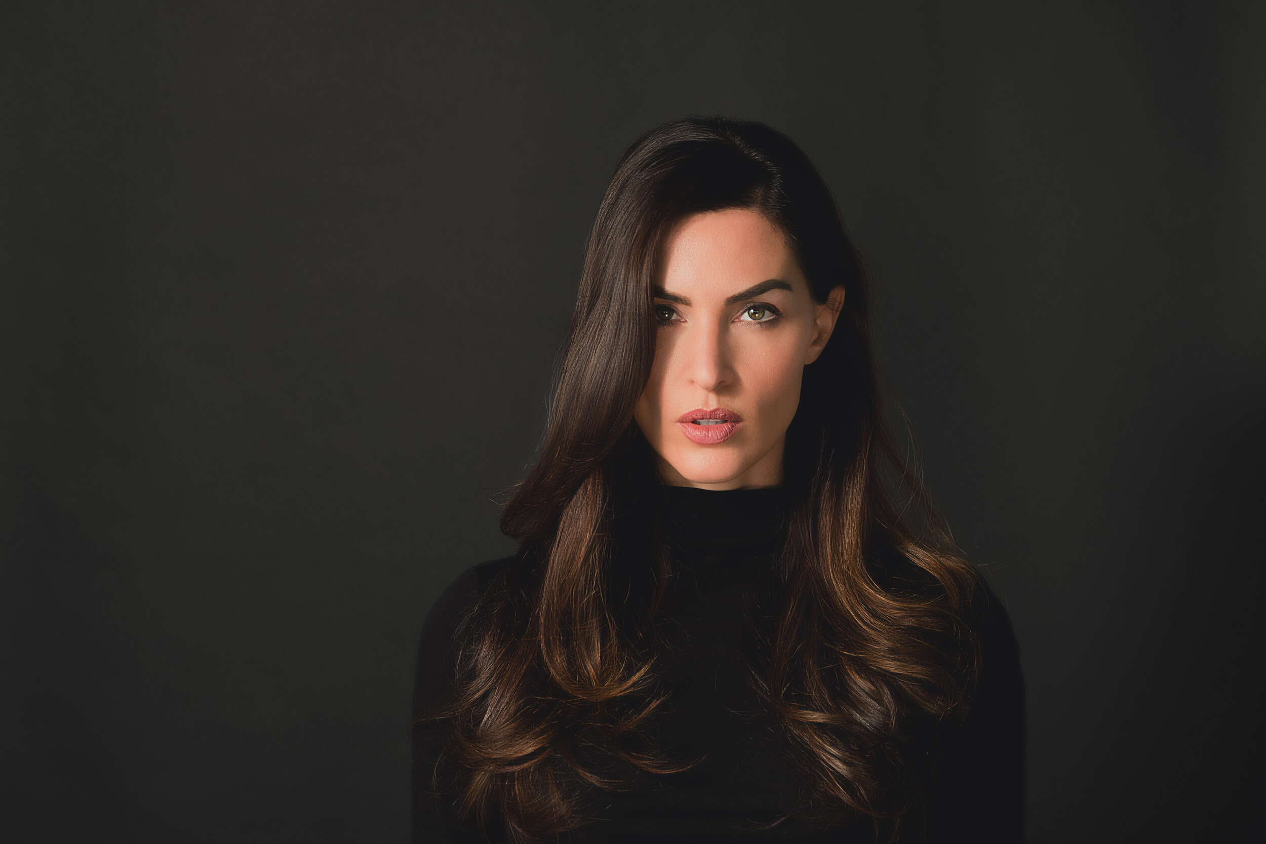 A woman with long brown hair and green eyes, wearing a black turtleneck, looking directly at the camera against a dark background.