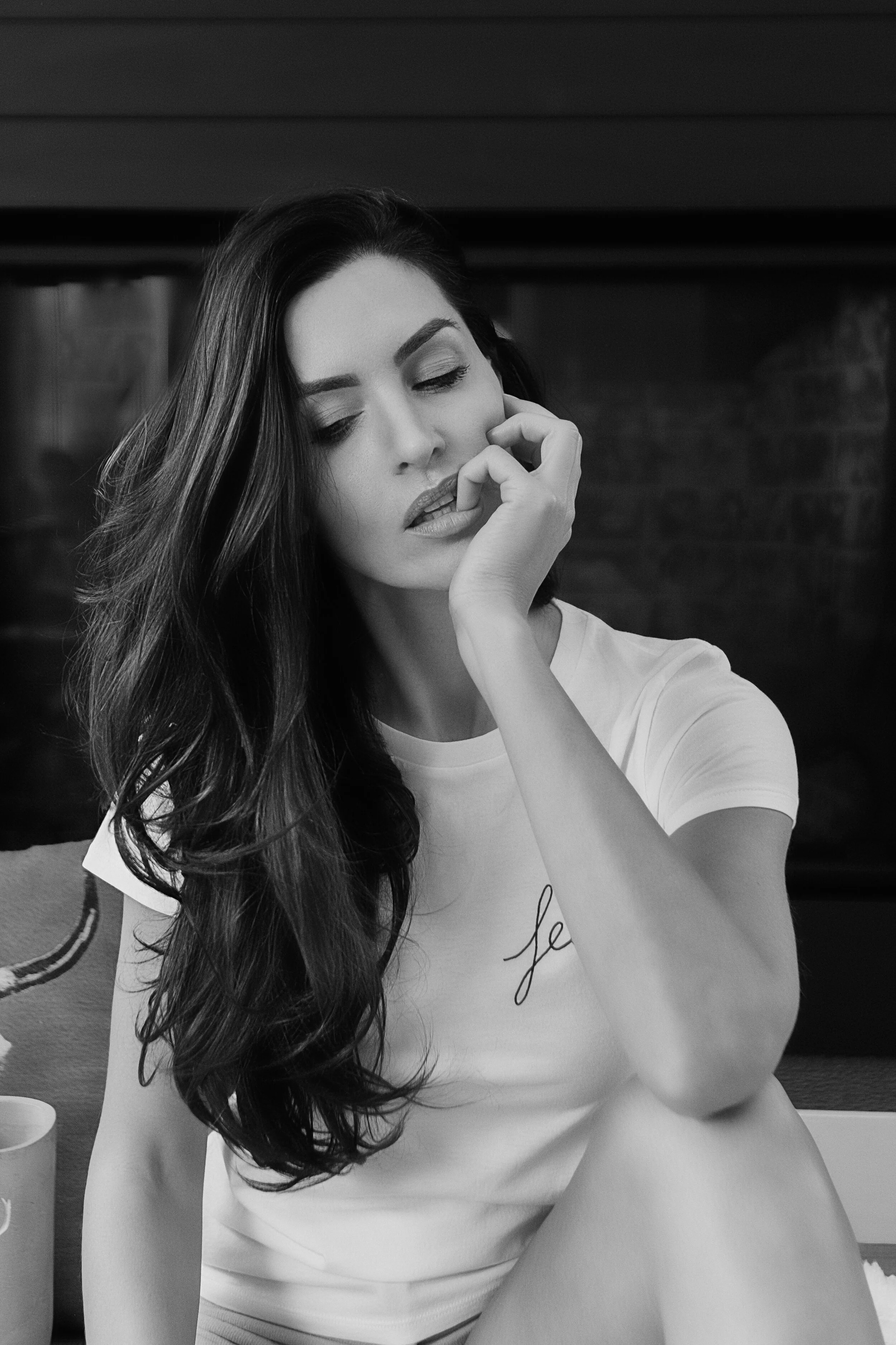A black-and-white photo of a woman with long dark hair, wearing a white t-shirt, sitting with her eyes closed, touching her face gently with her hand.