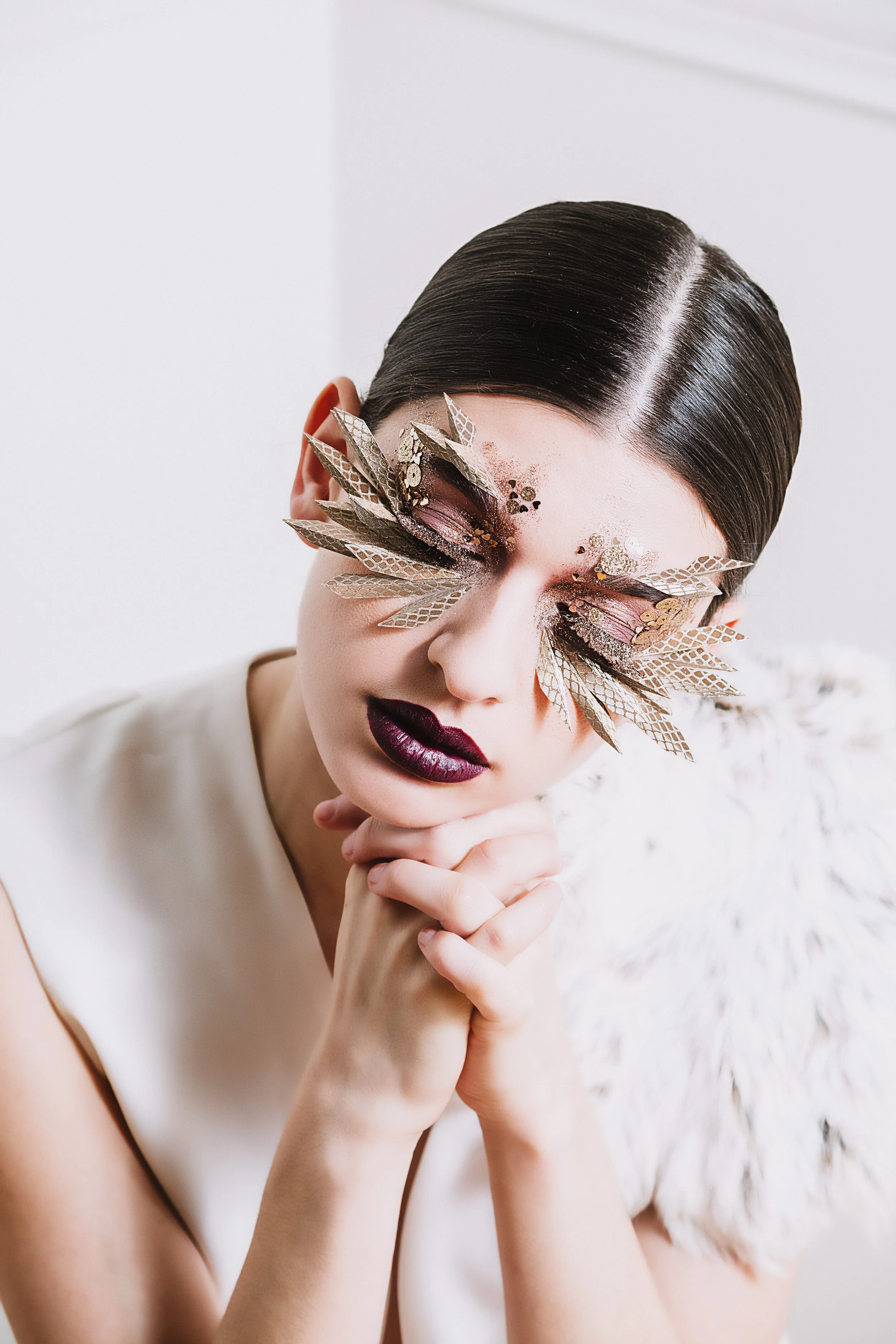 A woman with dark, slicked-back hair and dramatic dark lipstick wearing makeup with gold and pink eye decorations, resting her chin on her hands.
