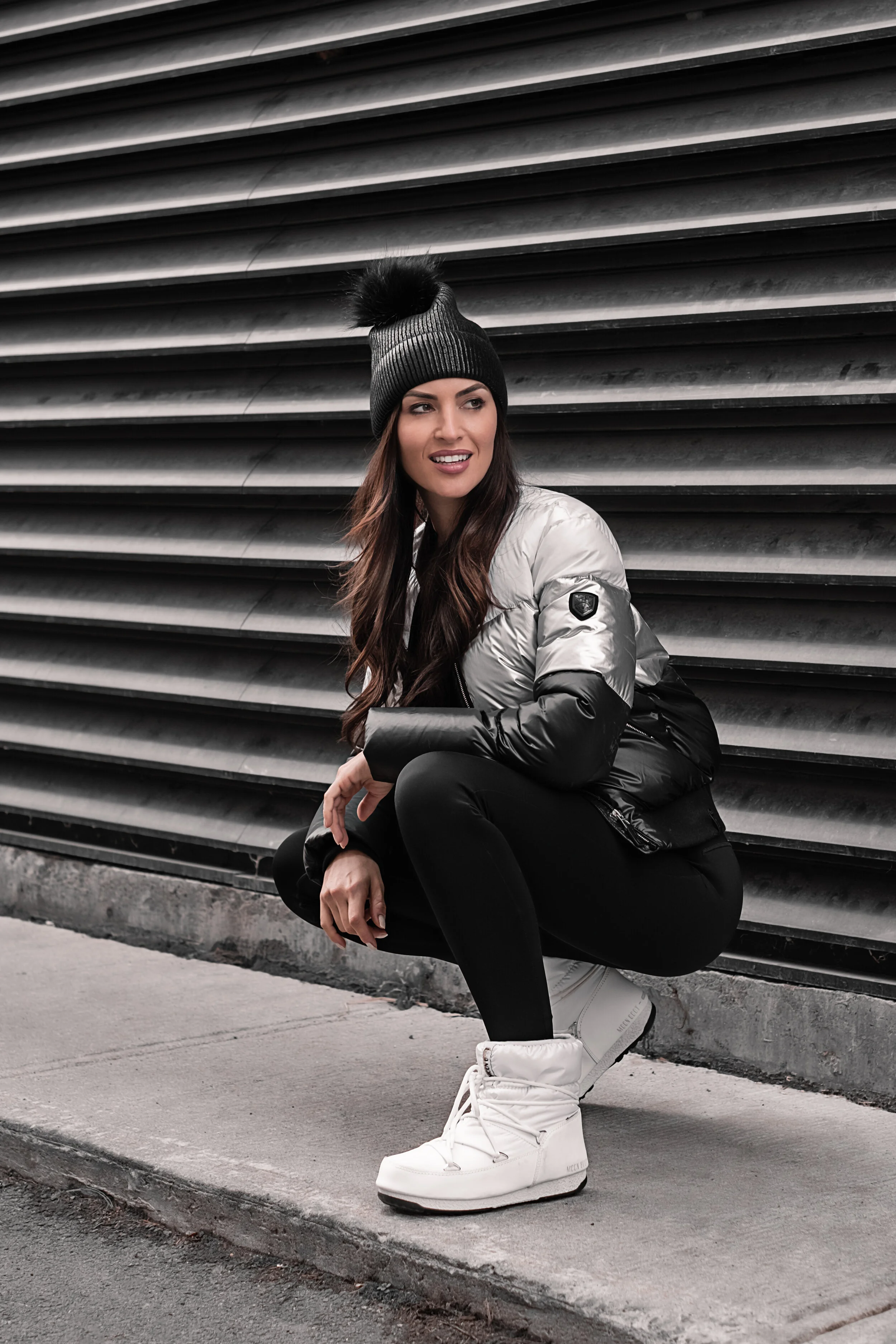 A woman in a black knit beanie, silver and black puffer jacket, black leggings, and white winter boots crouches on a sidewalk in front of a large vented metal wall.