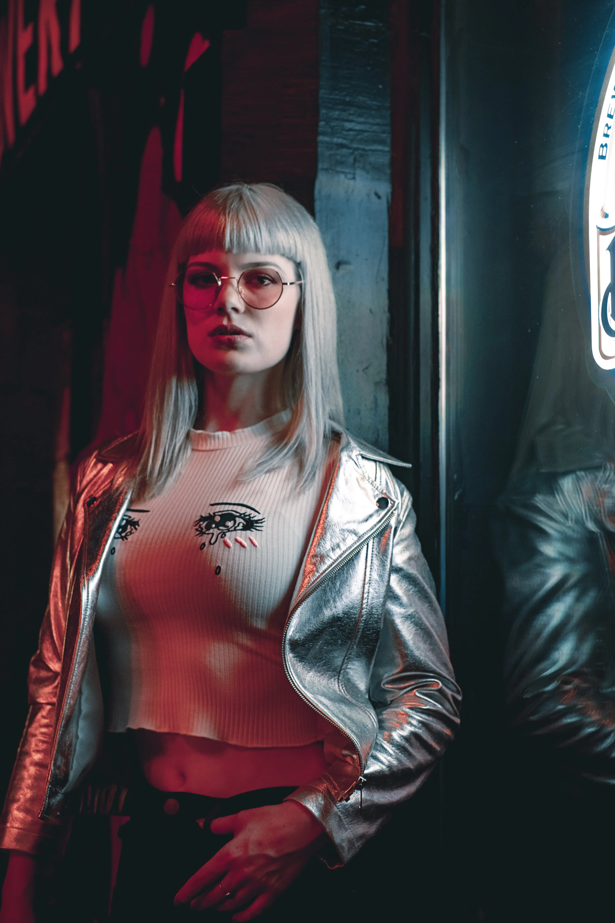 A young woman with platinum blonde hair in a bob cut and round glasses, wearing a white crop top with a graphic eye design and a metallic silver jacket, standing in front of a dark background illuminated by neon lighting.