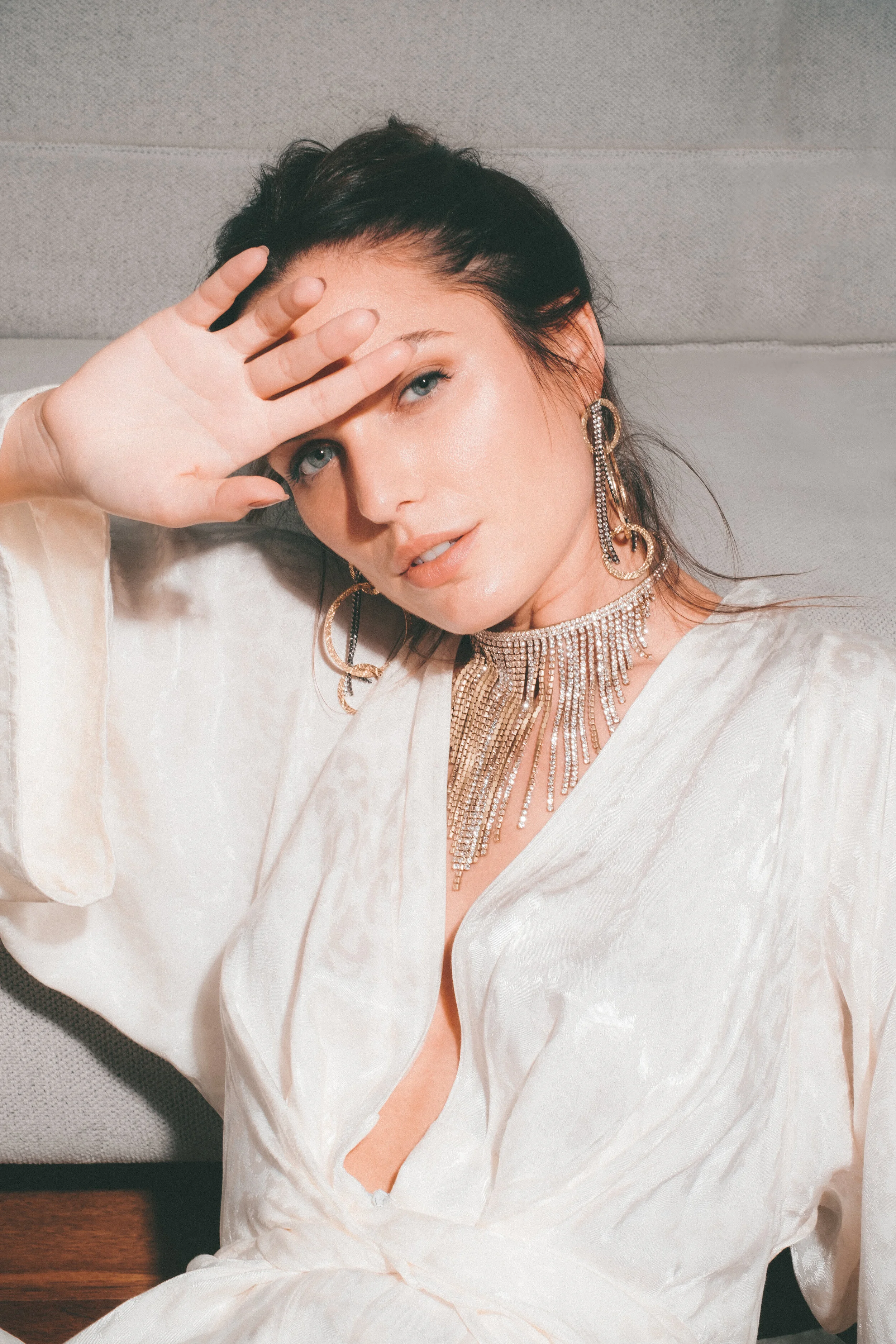A woman with dark hair and blue eyes, wearing a white silk robe, large pearl earrings, a sparkling rhinestone choker, and a matching necklace, lying on a bed with a gray headboard, holding her hand up to her forehead, looking at the camera.