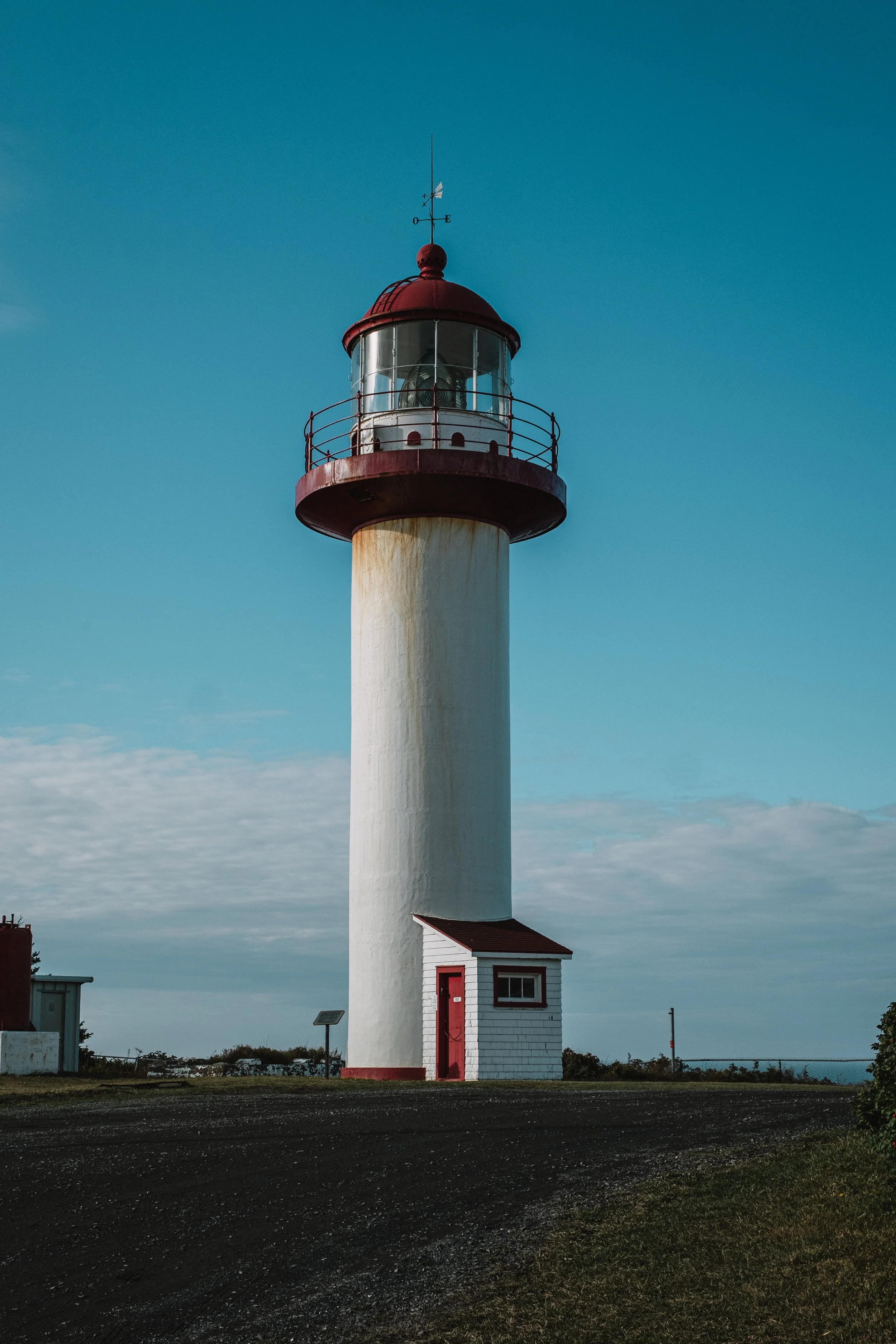 A lighthouse with a white cylindrical tower, red balcony, and red dome, standing against a partly cloudy sky.