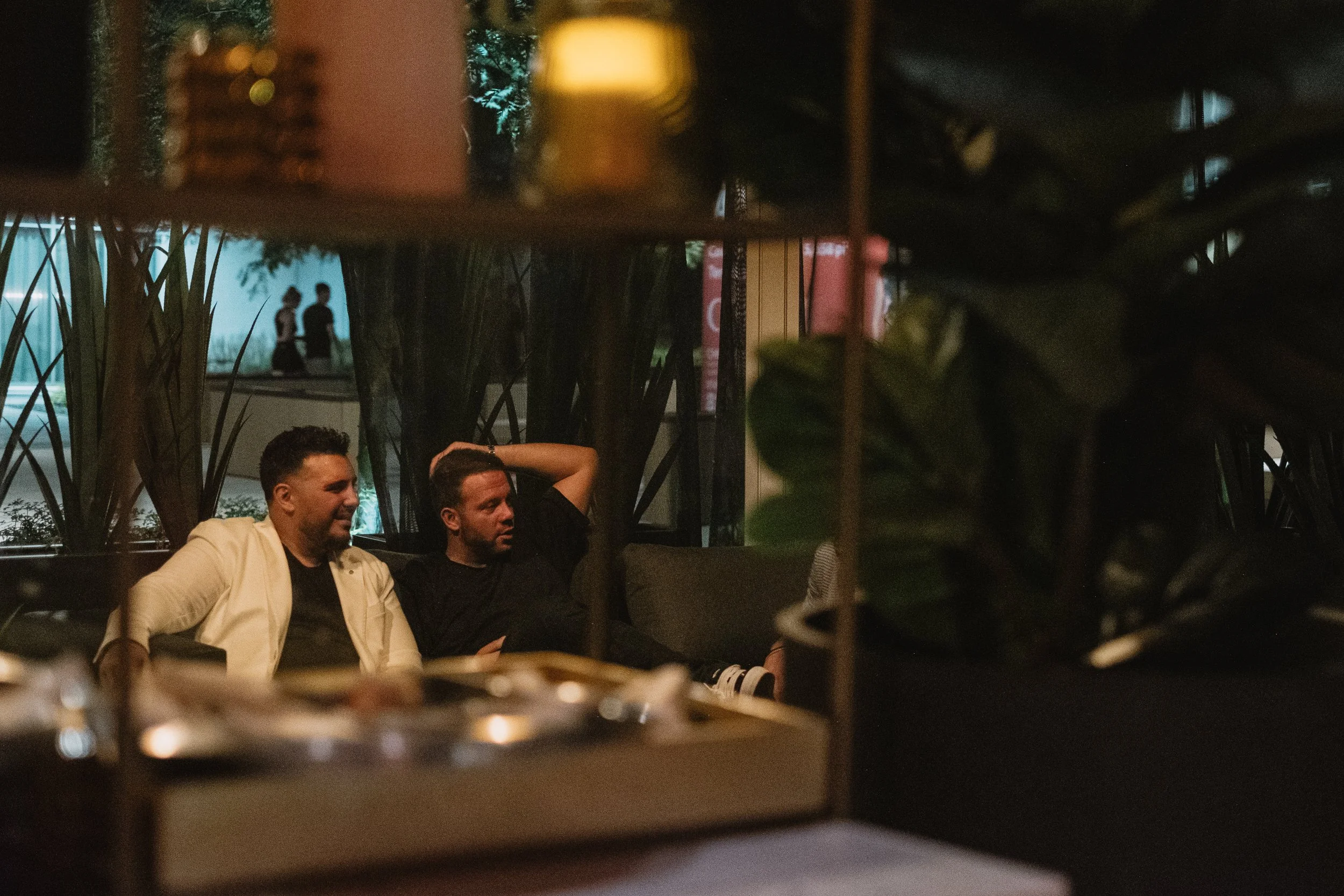 Two men sitting on a couch at night, one wearing a white jacket and the other in a black shirt, seen through a decorative partition with greenery in the foreground and silhouettes of two people walking outside in the background.