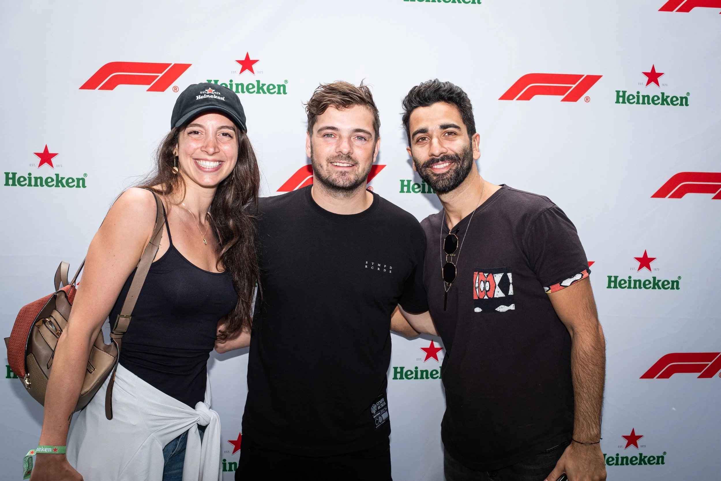 Three people smiling and standing close together in front of a backdrop with Heineken and F1 logos, at a daytime outdoor event.