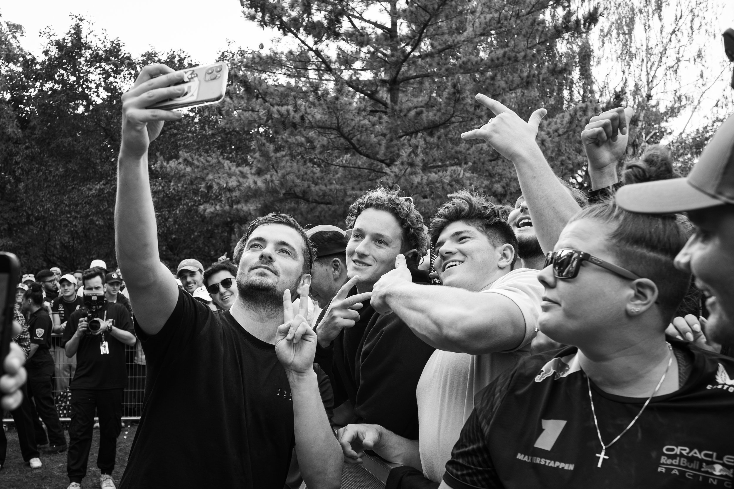 A group of smiling young men taking a selfie together outdoors, with others in the background and trees behind them.