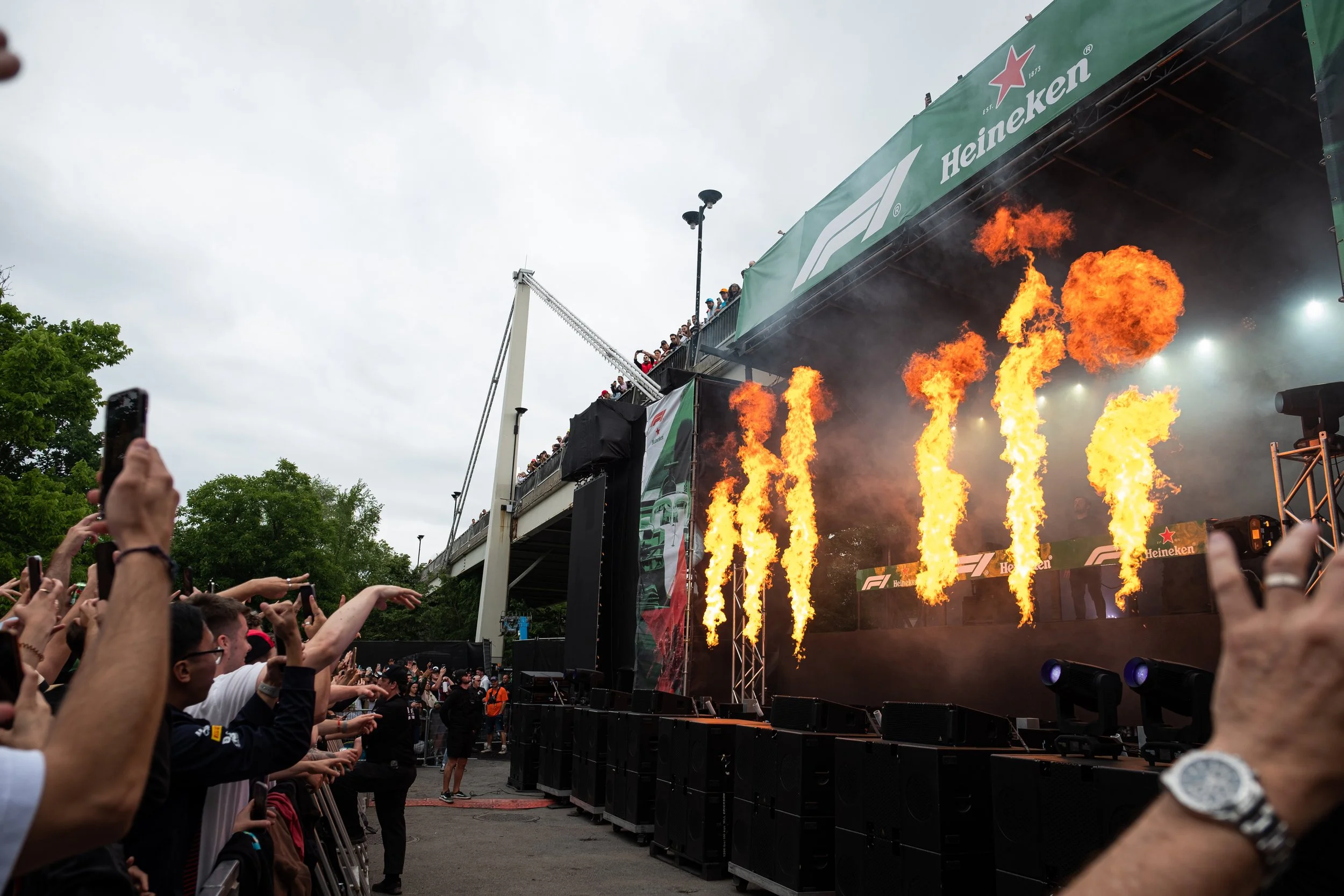 Audience watching a stage with flames at a motorsport event, large crowd on a balcony above, Heineken banners visible.