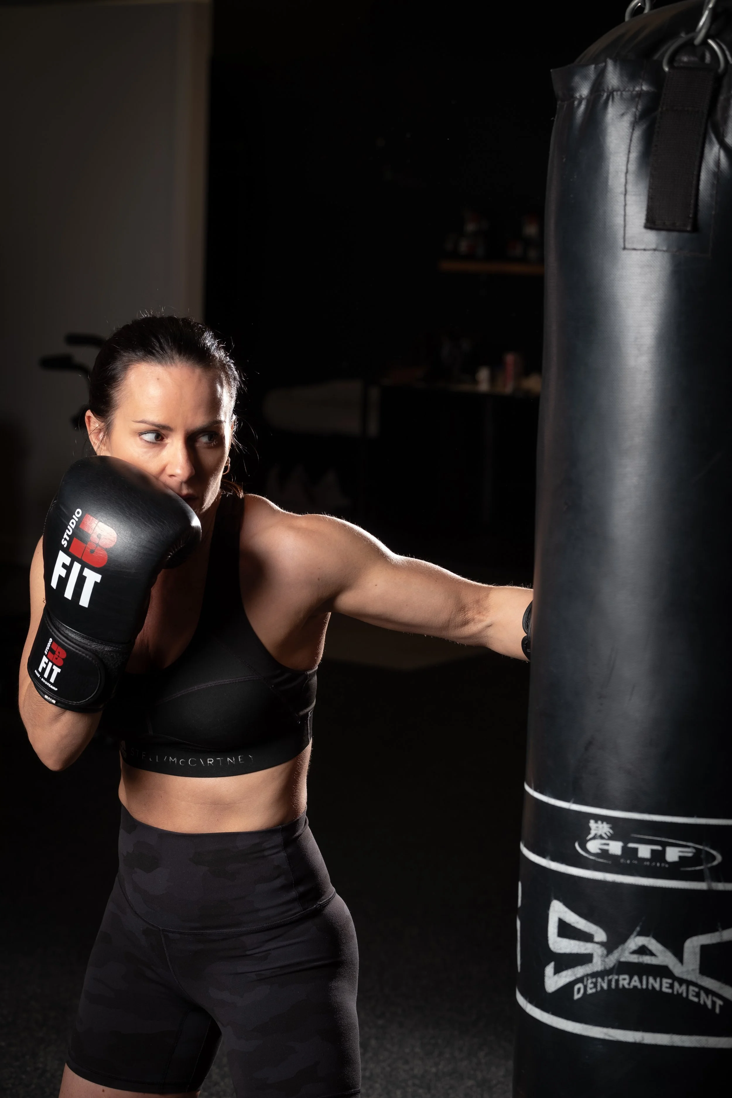 A woman with dark hair in a ponytail wearing black workout gear and boxing gloves punches a black punching bag in a gym.