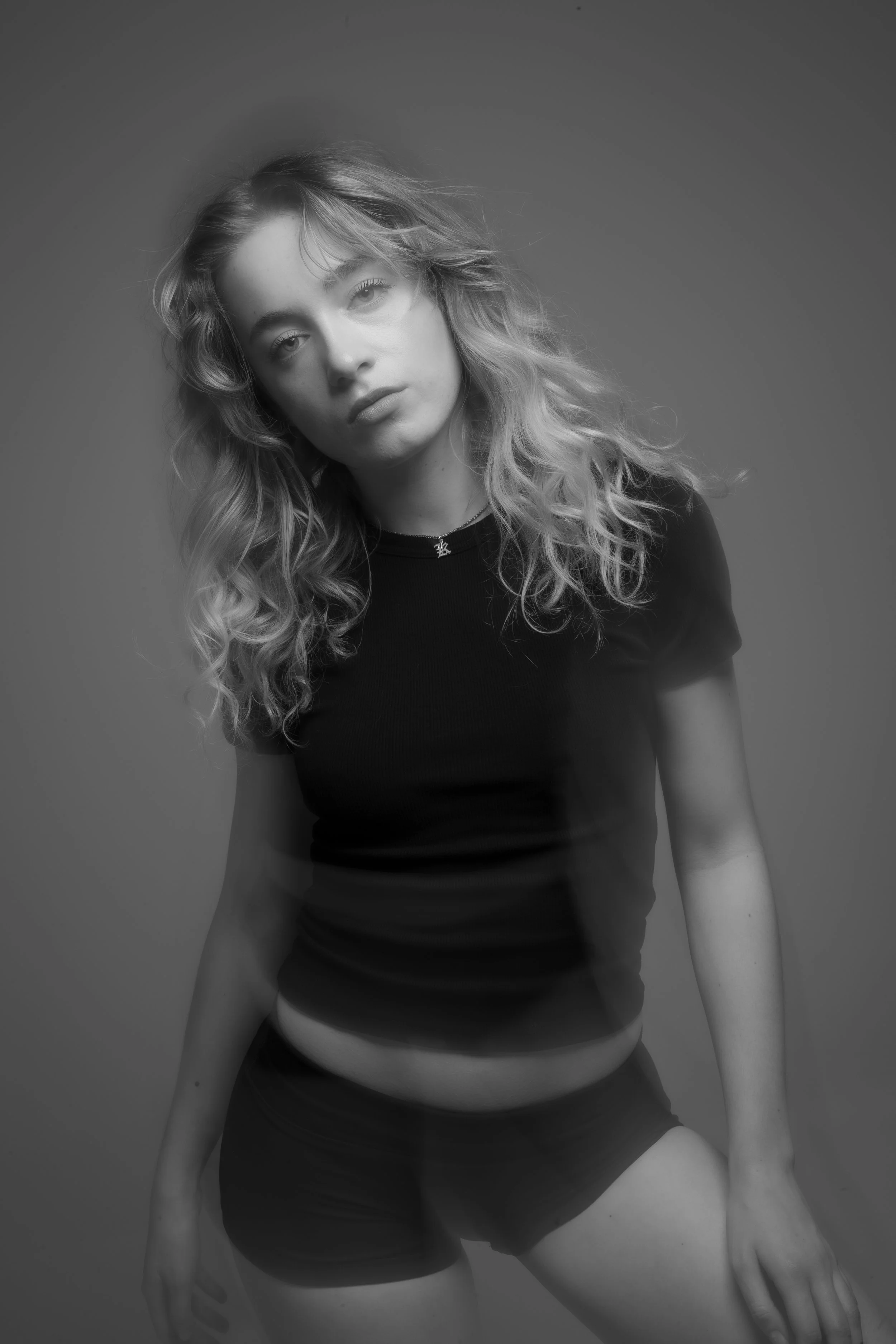 Black and white portrait of a young woman with curly hair, wearing a black t-shirt and shorts, looking directly at the camera.