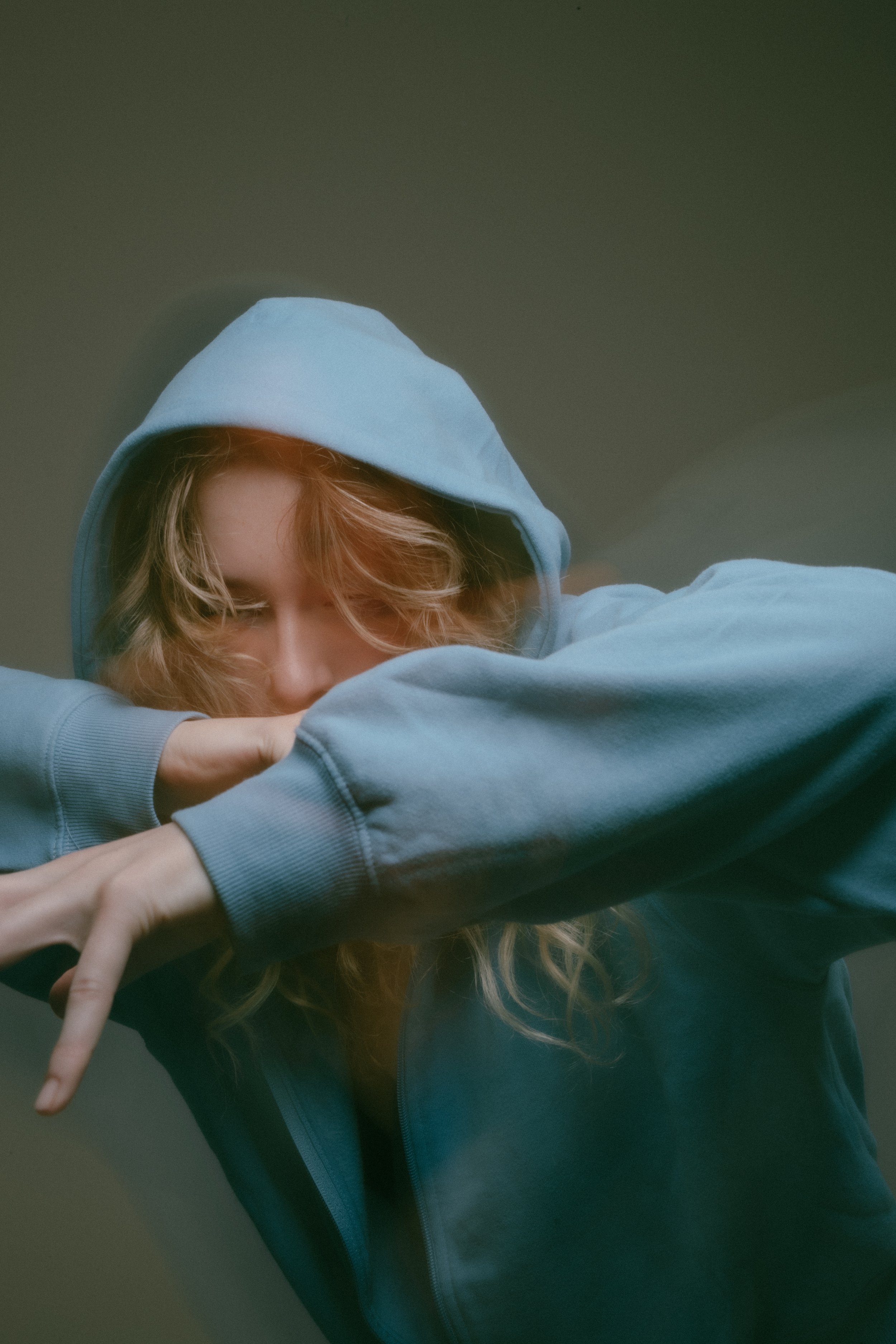 A young woman with wavy red hair wearing a light blue hoodie with the hood up, resting her face on her forearm with her eyes closed.